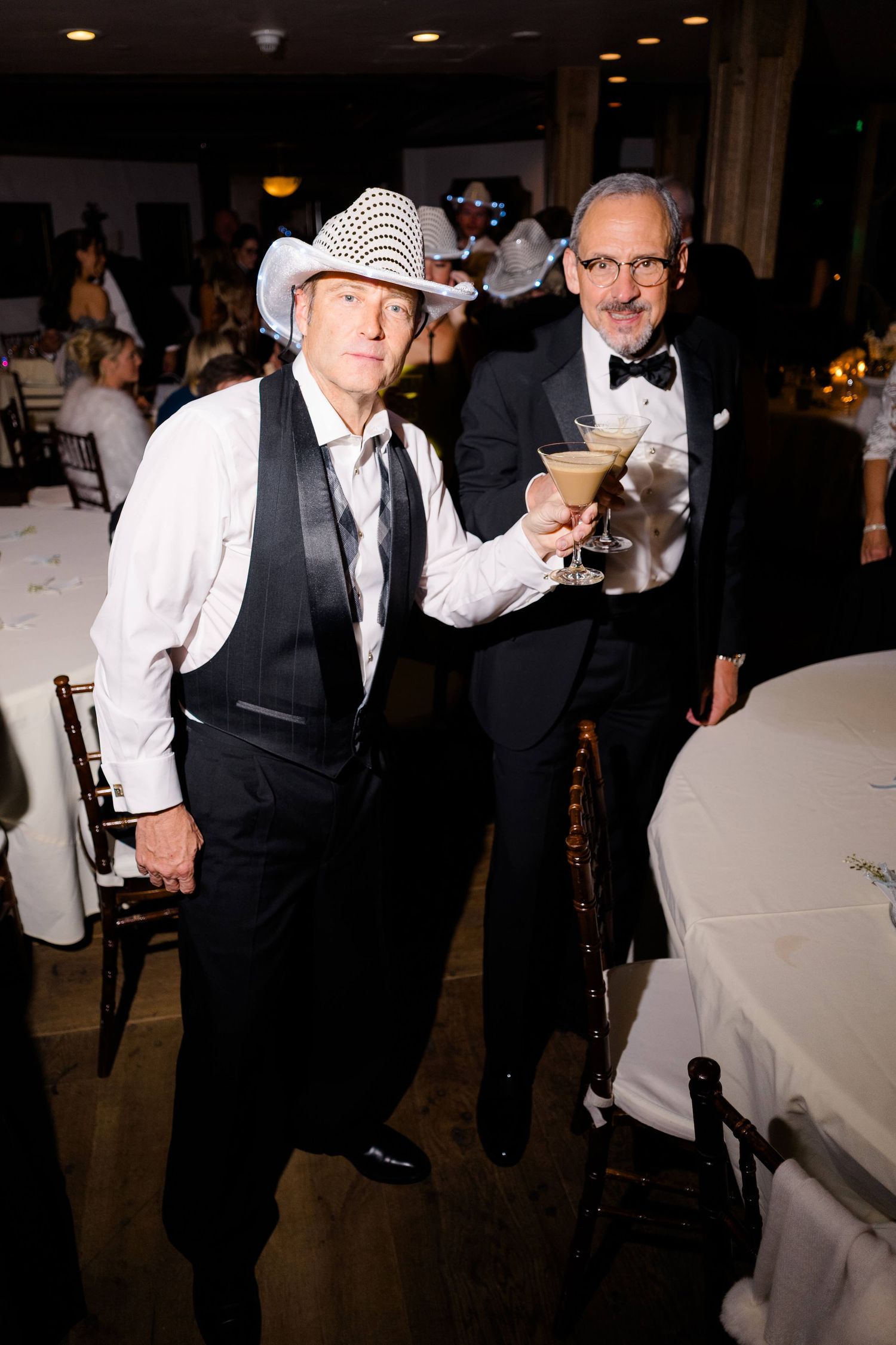 Two men in formal attire pose at an elegant dinner event, one wearing a white fedora and the other holding a martini glass.