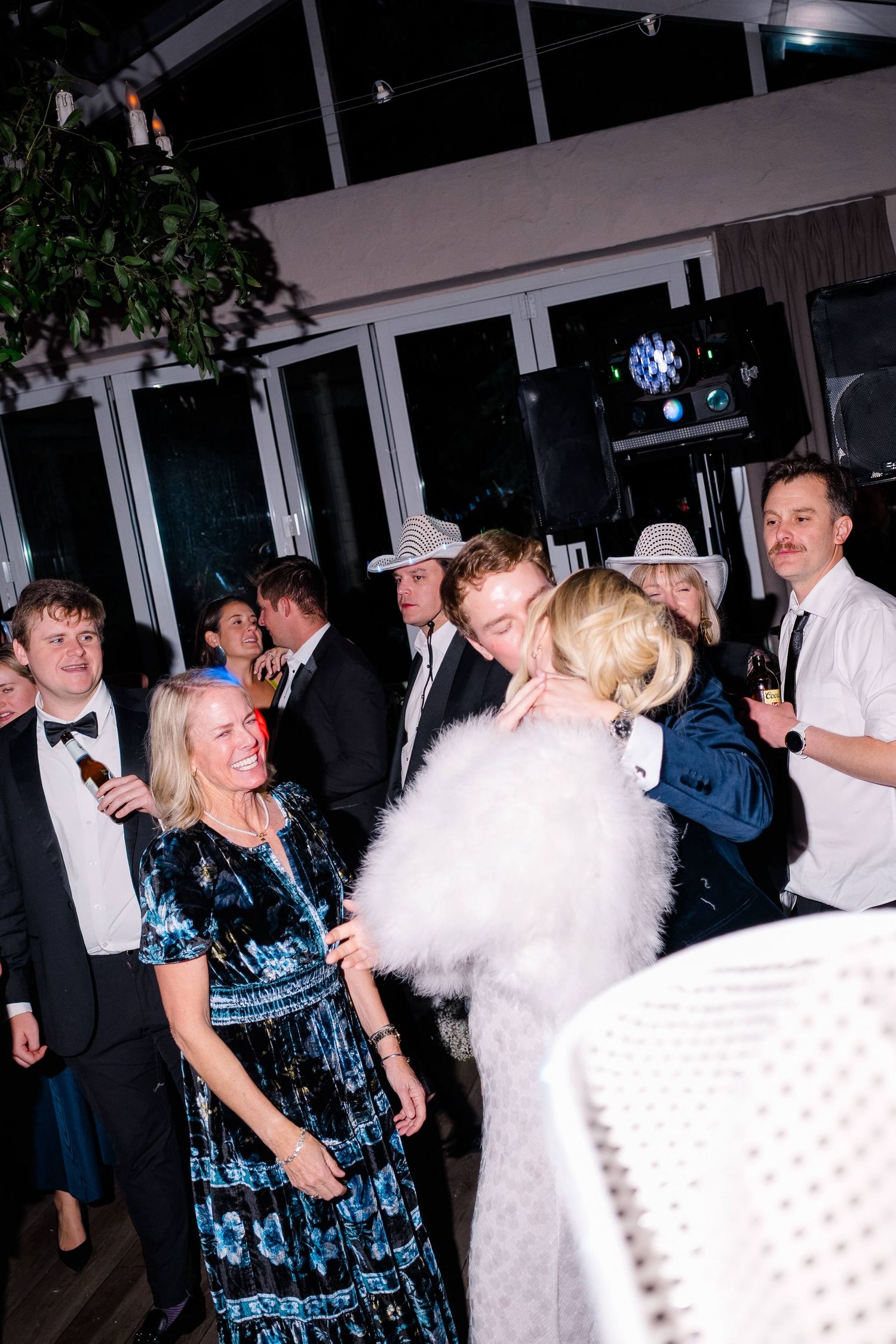 Guests in formal attire dance and embrace at an elegant evening party with festive lighting.
