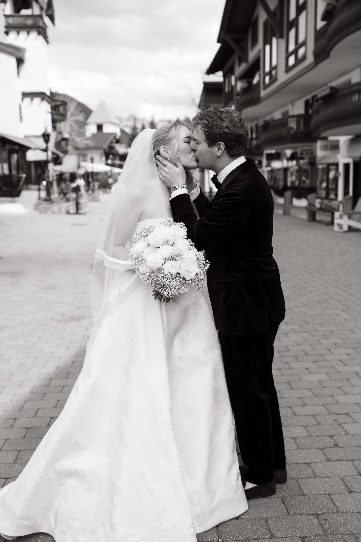 Black and white photo of a bride and groom sharing a kiss outdoors, bride holding a bouquet in a elegant white gown.