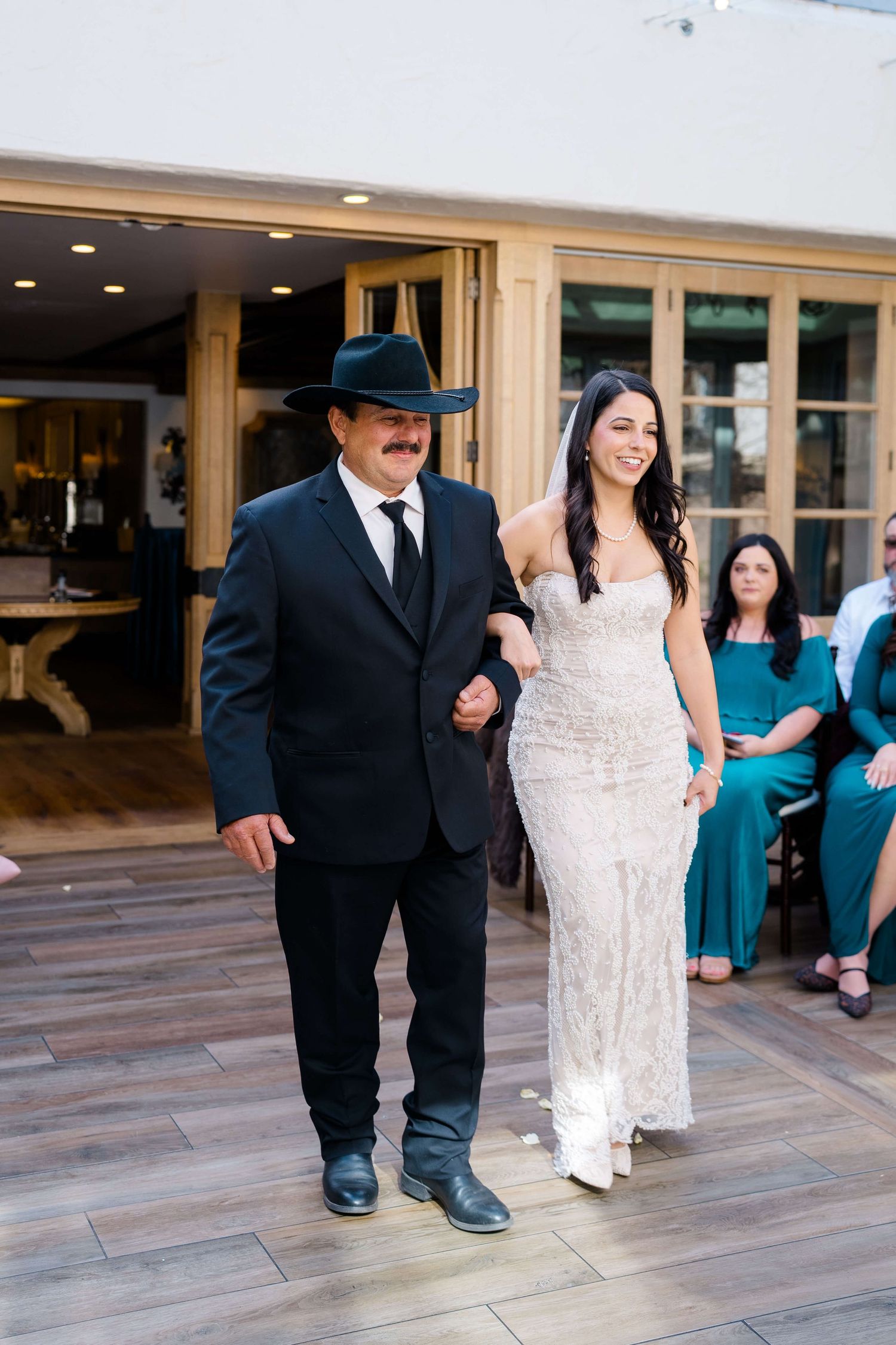 A bride in a white lace gown walks arm-in-arm with her father in a black suit and cowboy hat at a wedding reception.