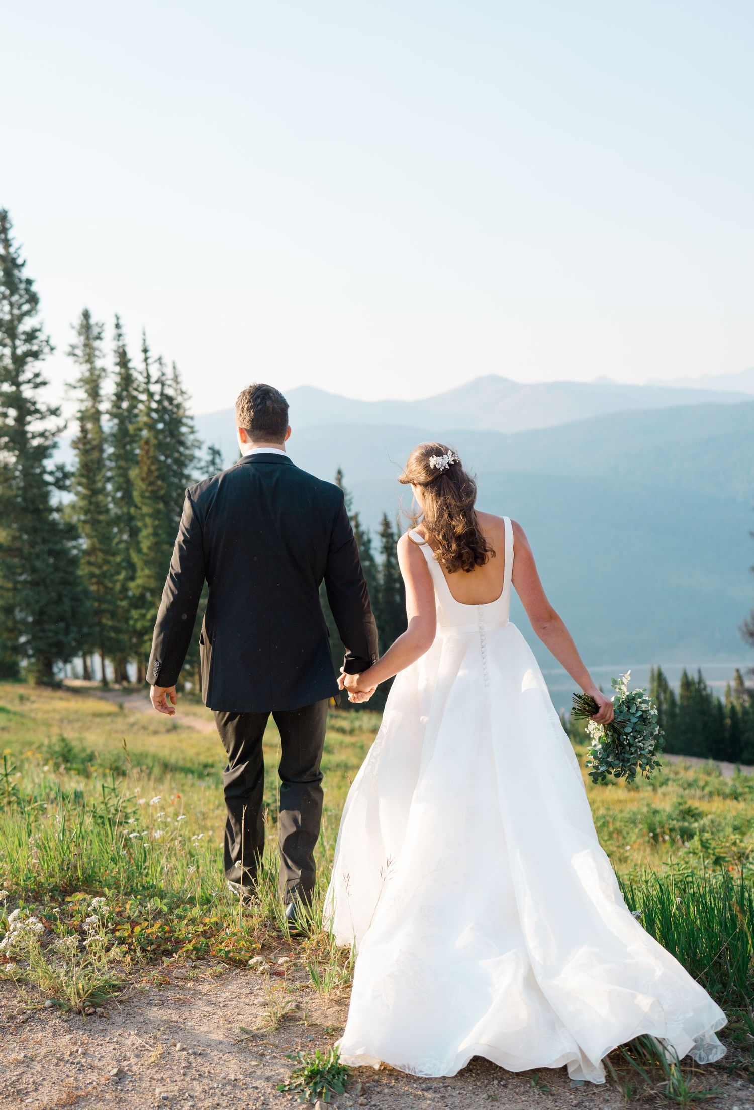 A couple in wedding attire walks along a mountain trail with evergreen trees and scenic mountain views in the background at copper mountain.