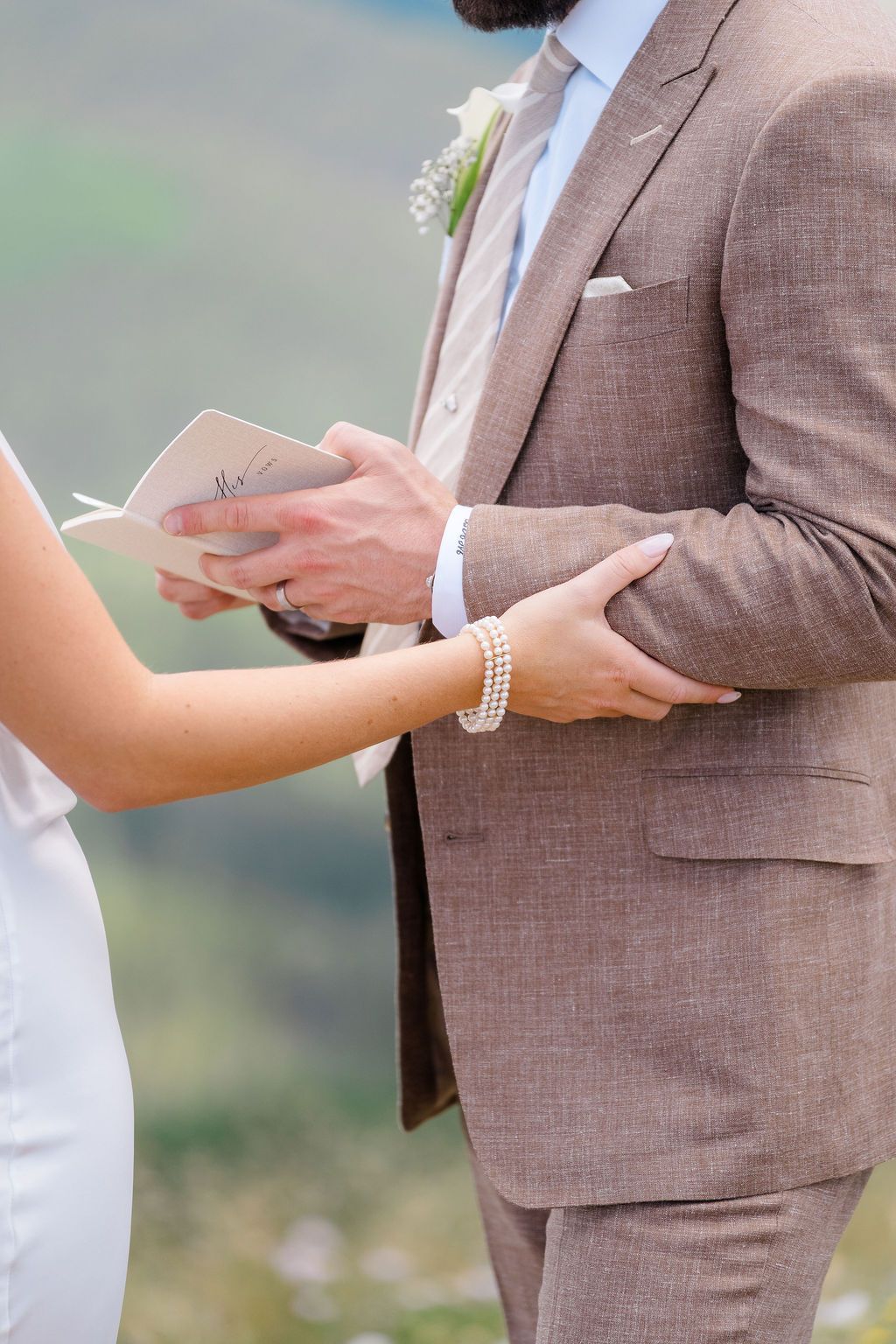Bride in pearl bracelet reads wedding vows from booklet while groom in brown suit holds her hand during outdoor ceremony.