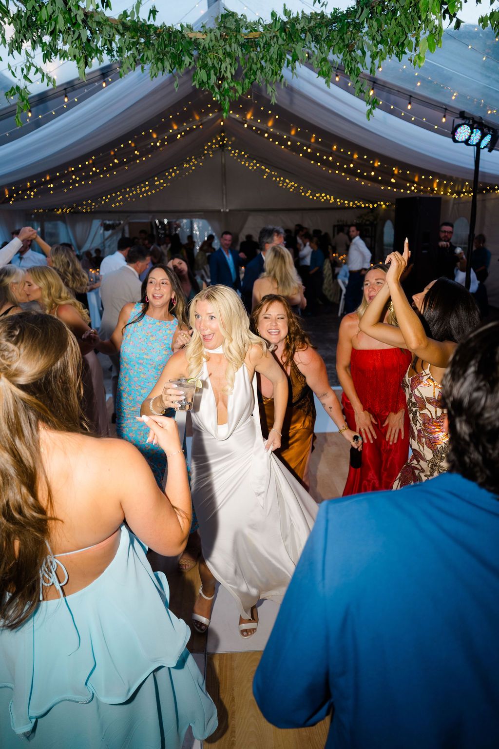 Guests dancing joyfully at an outdoor wedding reception under a tent with string lights.