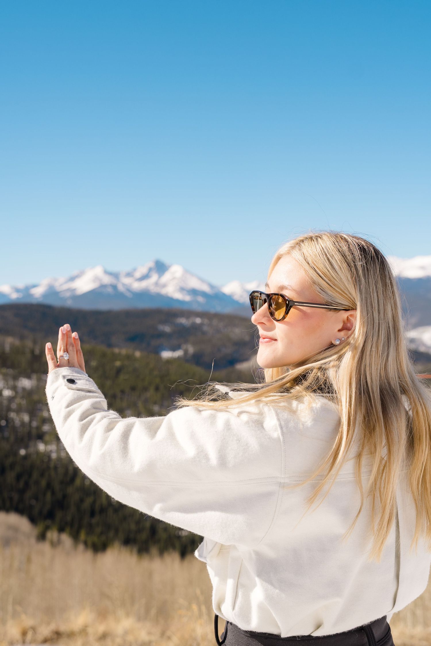 Blonde woman in sunglasses and white jacket posing with hands raised against snow-capped mountain backdrop.