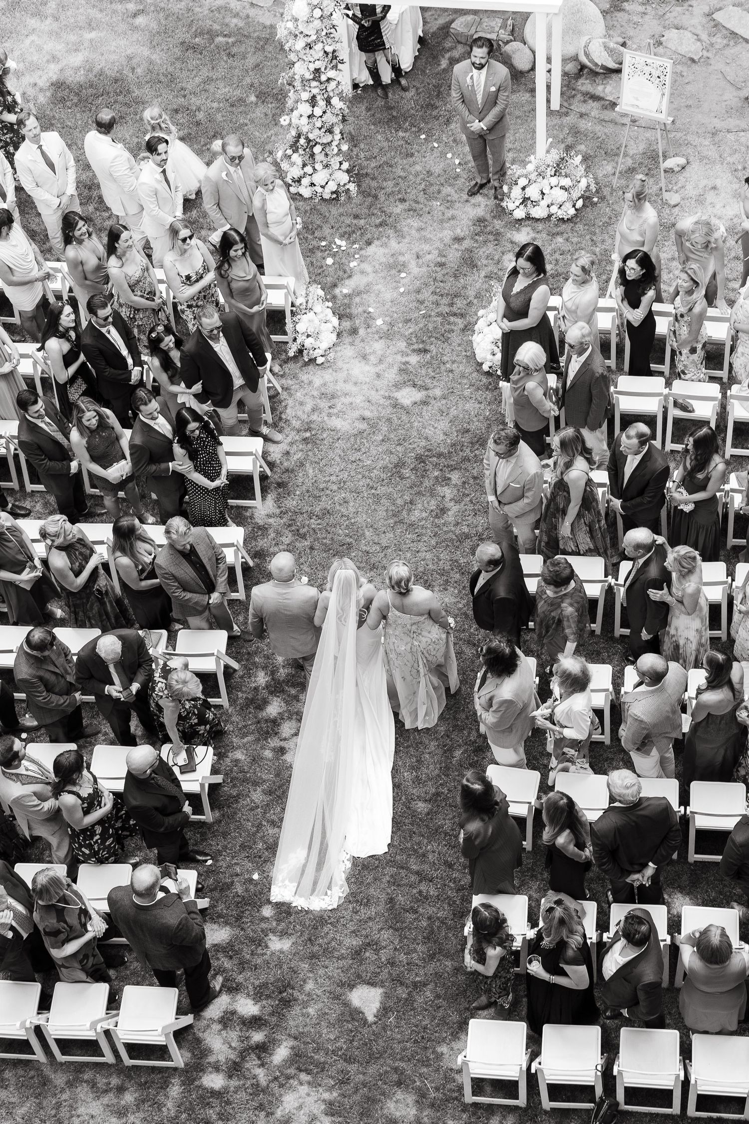 Aerial black and white photo of a bride walking down the aisle at an outdoor wedding ceremony.