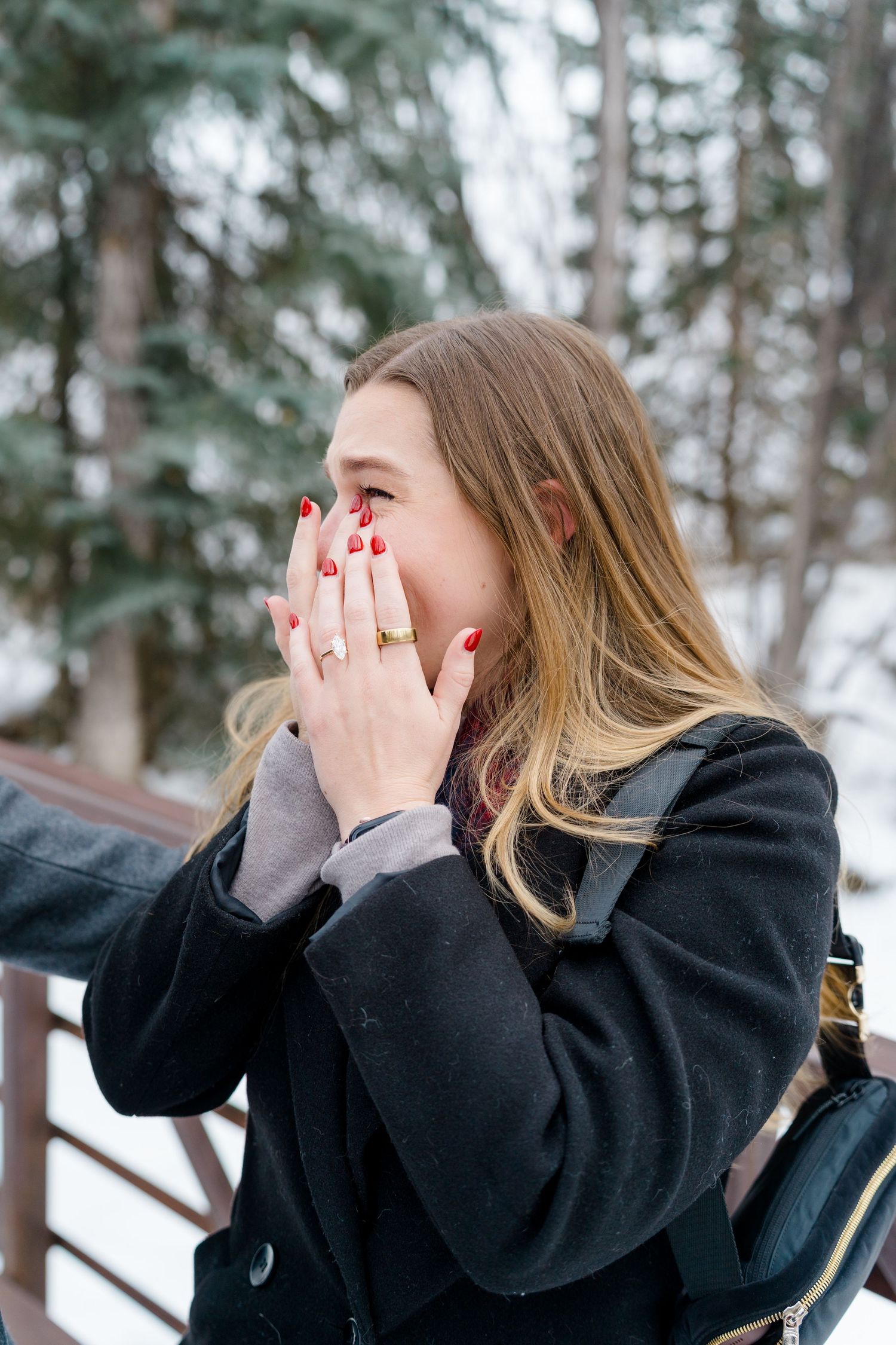 Person in black coat covers their face with hands while standing in snowy winter forest setting.