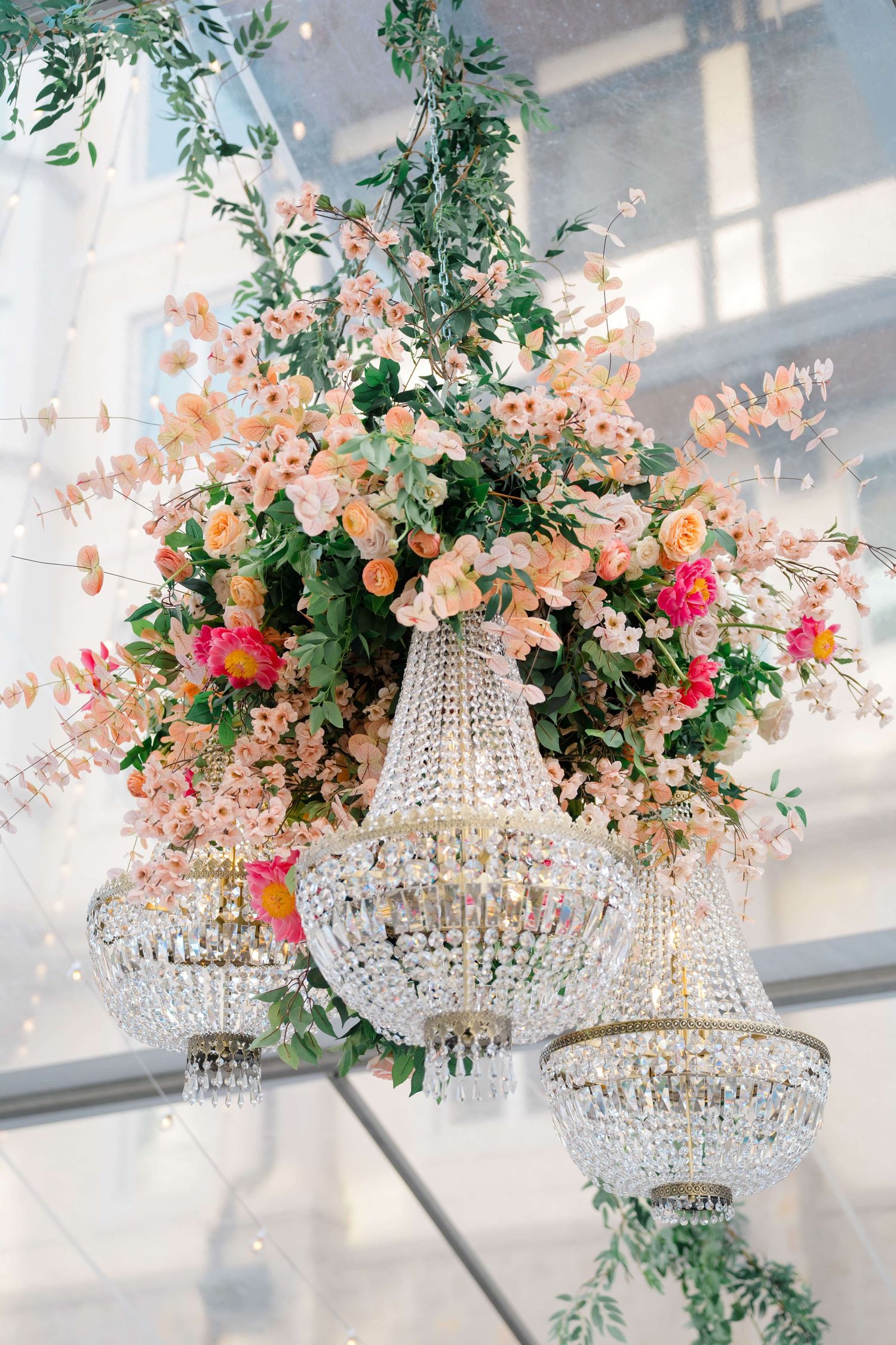 Arrabelle Vail Wedding - Crystal chandeliers adorned with lush peach and pink floral arrangements hang elegantly in a bright wedding venue.