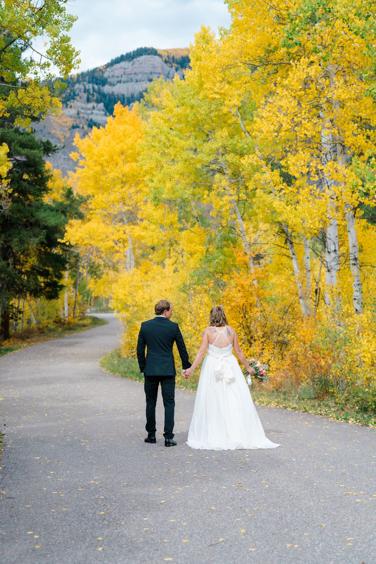 Bride and groom walking hand in hand on a winding path surrounded by vibrant yellow autumn aspen trees in the mountains.