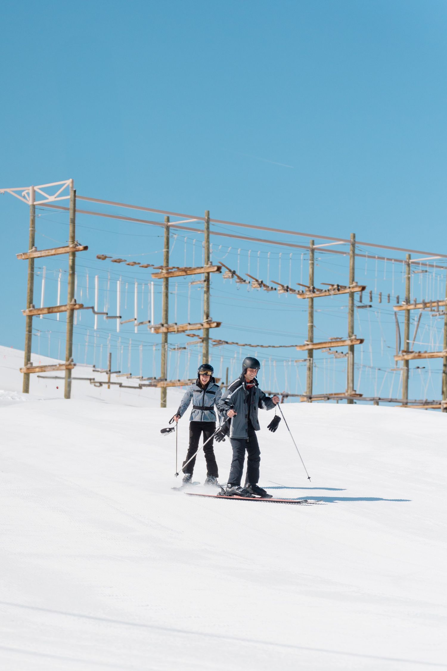 Skiers navigate through a snowy slope with wooden ski lift poles and cables visible against a bright blue sky.
