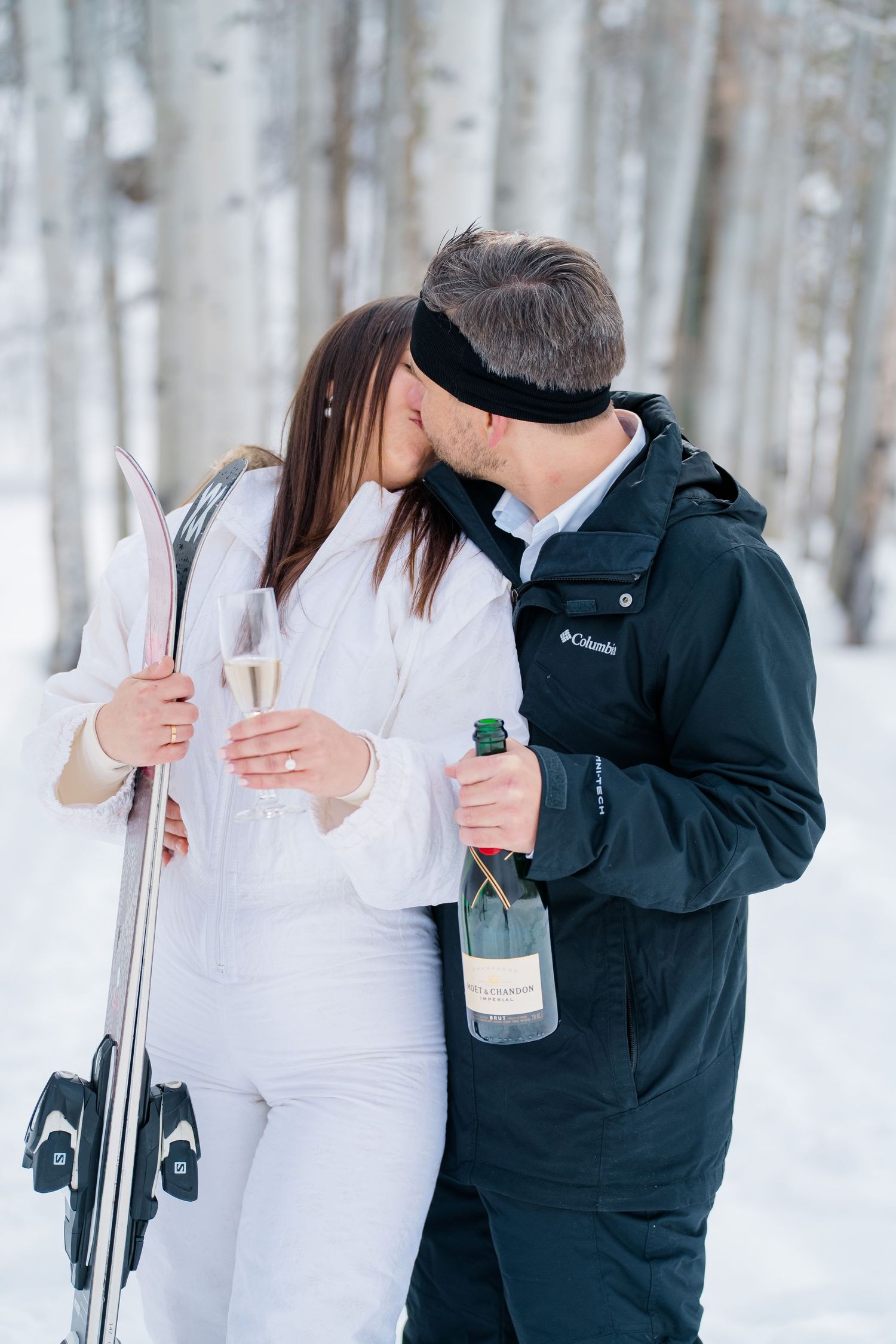 A couple in winter attire shares champagne while standing with skis in a snowy forest setting.