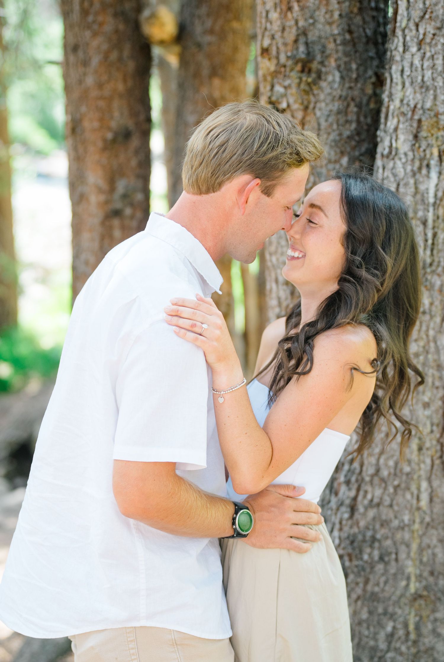 A romantic couple shares tender moments against tree trunks in a forest setting, wearing casual white and beige attire.
