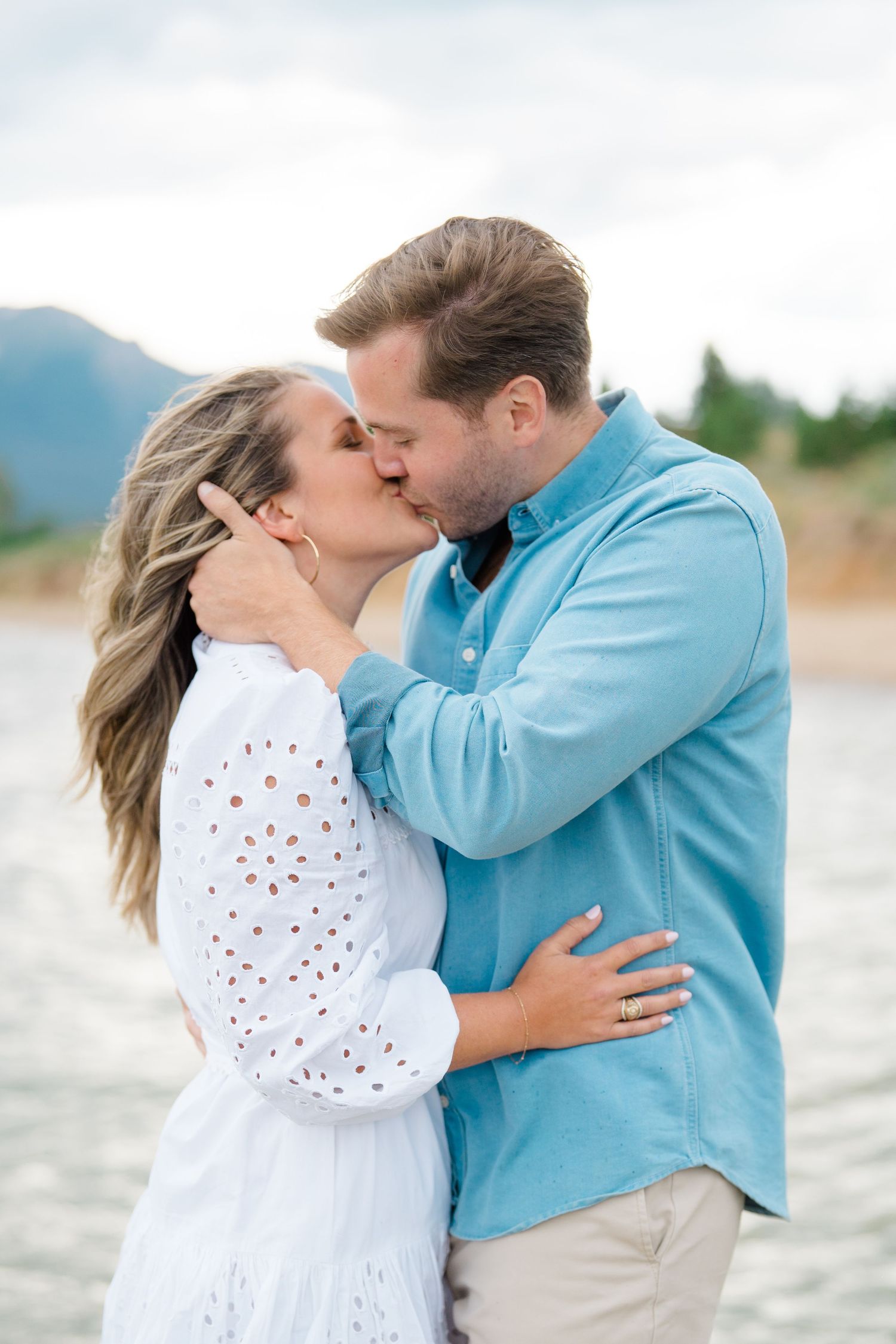 A romantic couple shares tender kisses on a beach wearing white and blue outfits with mountains in the background.