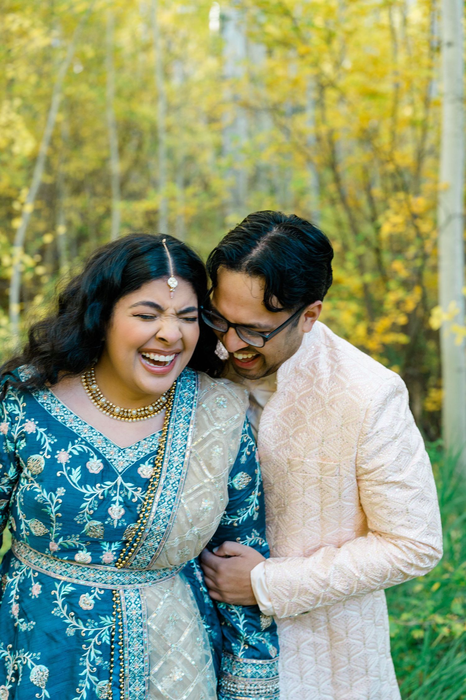 A couple in traditional South Asian wedding attire share a joyful moment against autumn foliage.