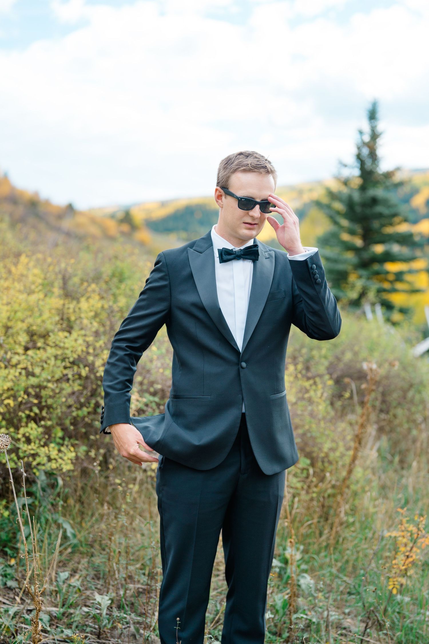 Groom in black tuxedo and bowtie poses playfully in outdoor field with autumn foliage.