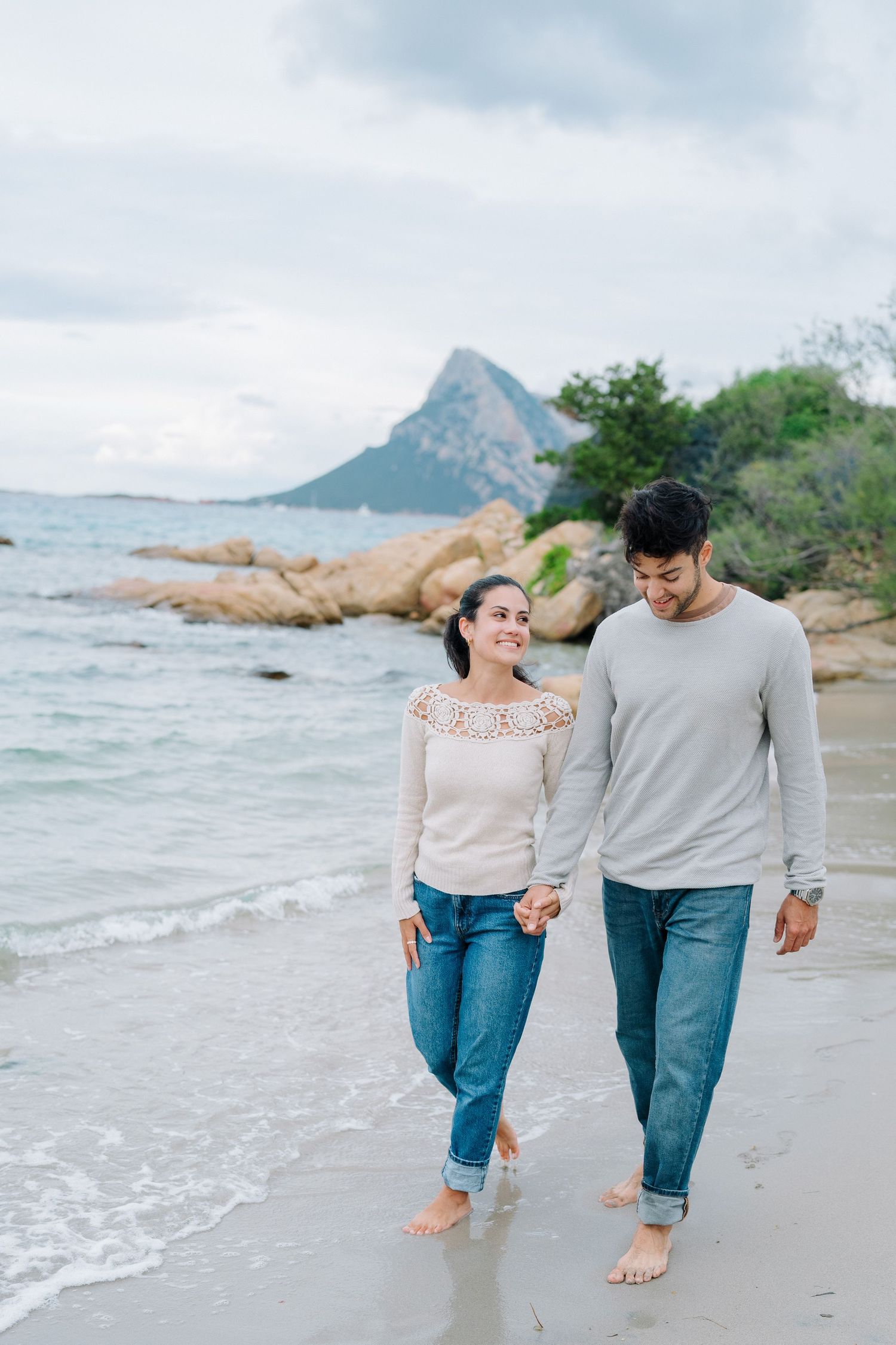 Couple walking barefoot along a beach with rocky outcrops and ocean waves in the background.