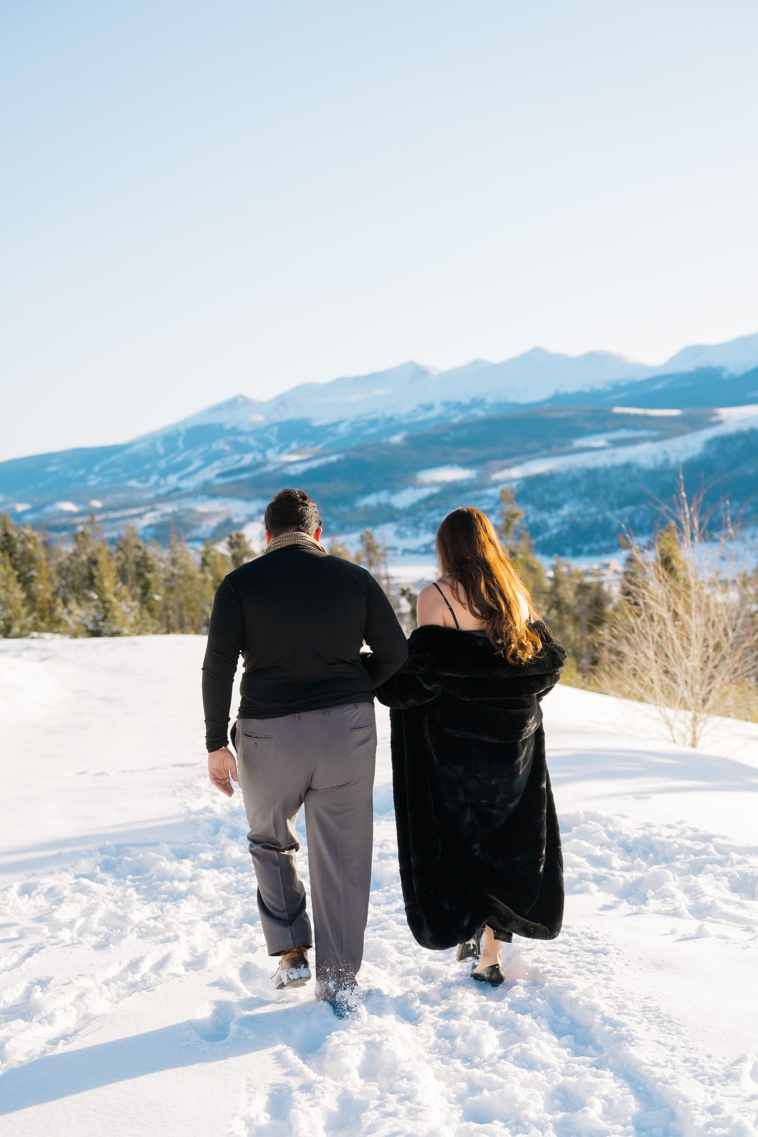A couple sits together in deep snow overlooking a scenic mountain landscape on a bright winter day.