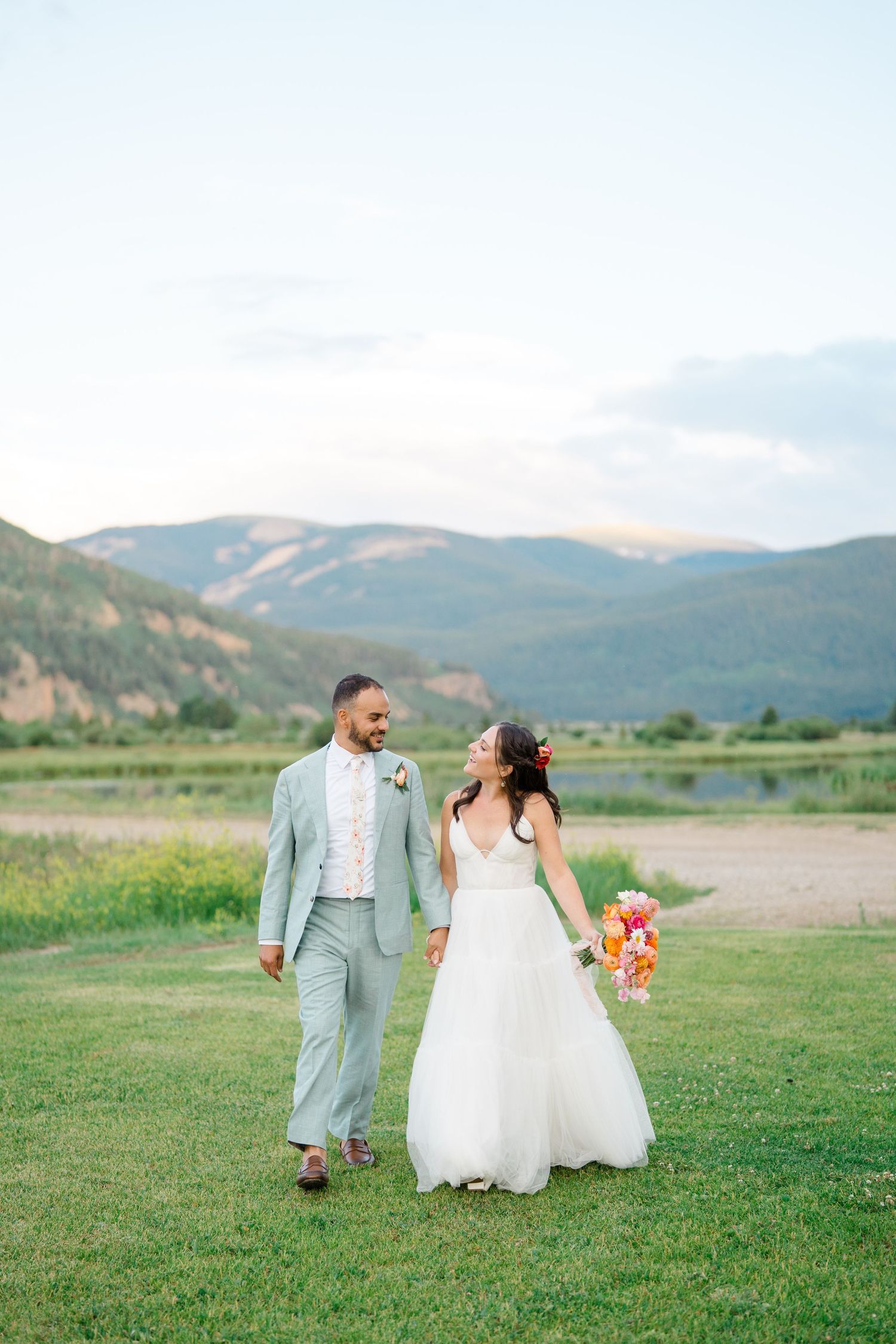 A wedding couple poses on grassy hills with mountains and lake views in the background.