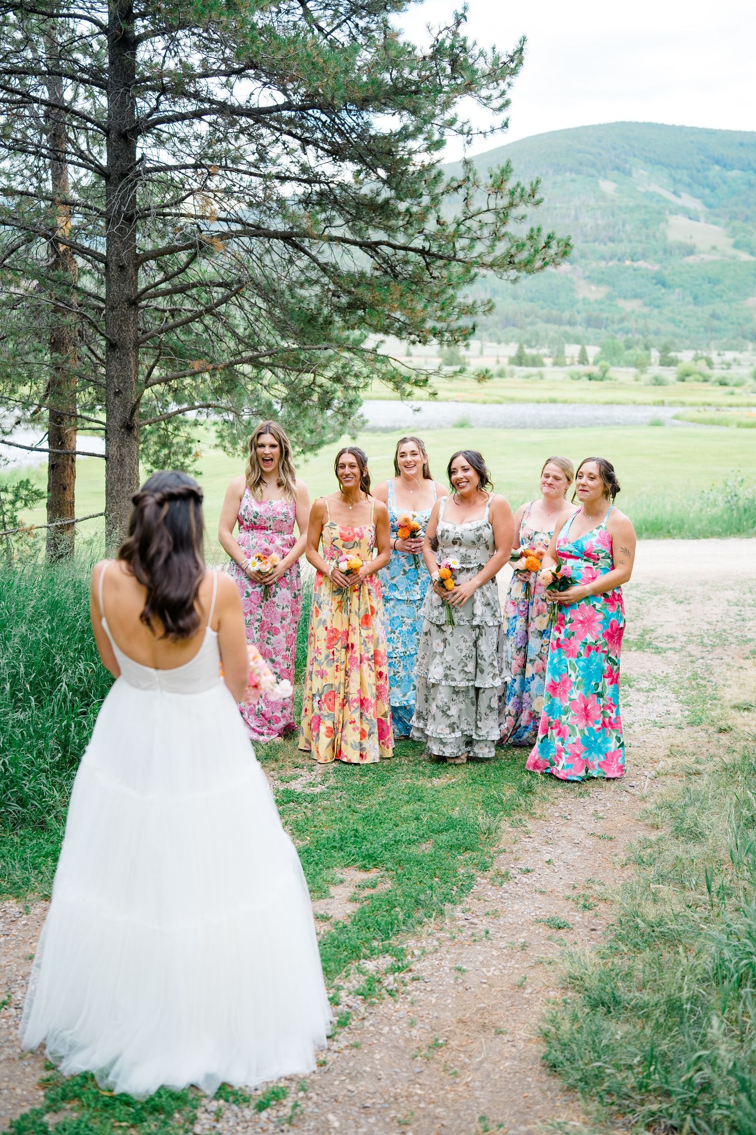 Wedding party in colorful floral dresses poses together in front of pine trees with mountain backdrop.