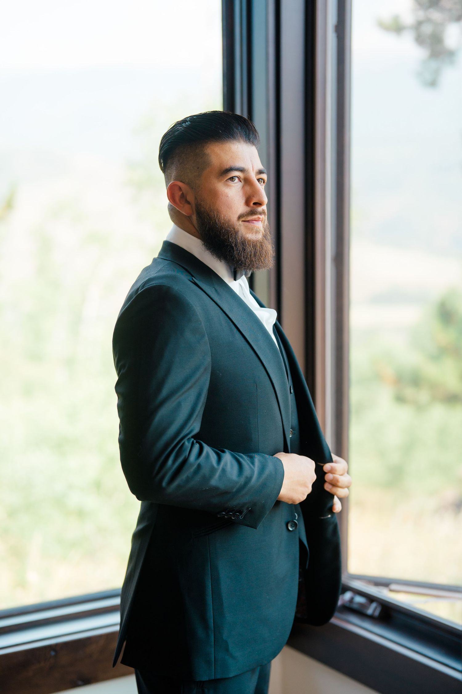 A bearded man in a dark suit stands thoughtfully by large windows in an elegant indoor setting.