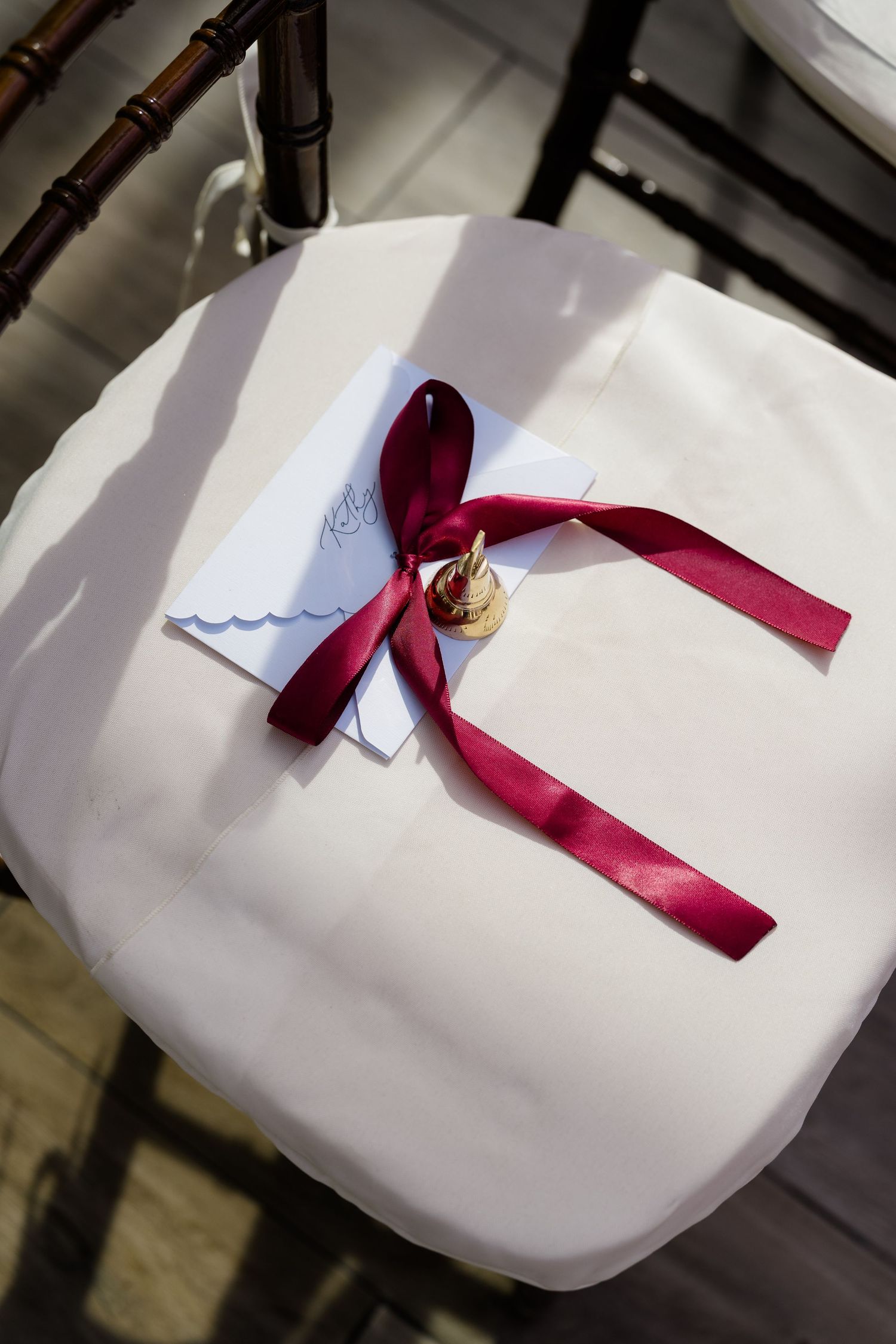 Elegant wedding chair with white cushion, burgundy ribbon bow, gold bell, and handwritten note card decoration.