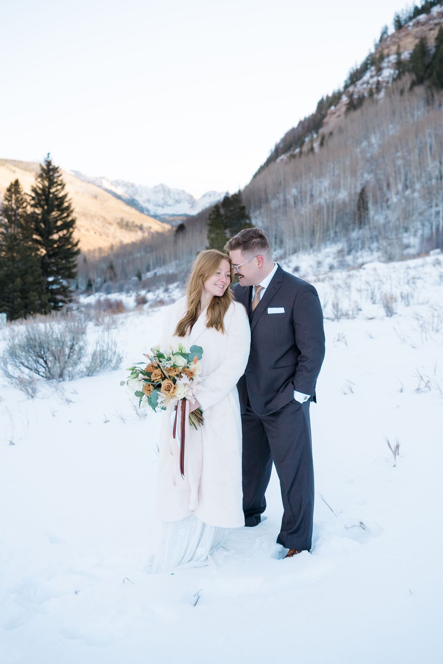 Bride and groom embrace in snowy mountain landscape, she holds autumn bouquet, wearing white coat, he in dark suit.