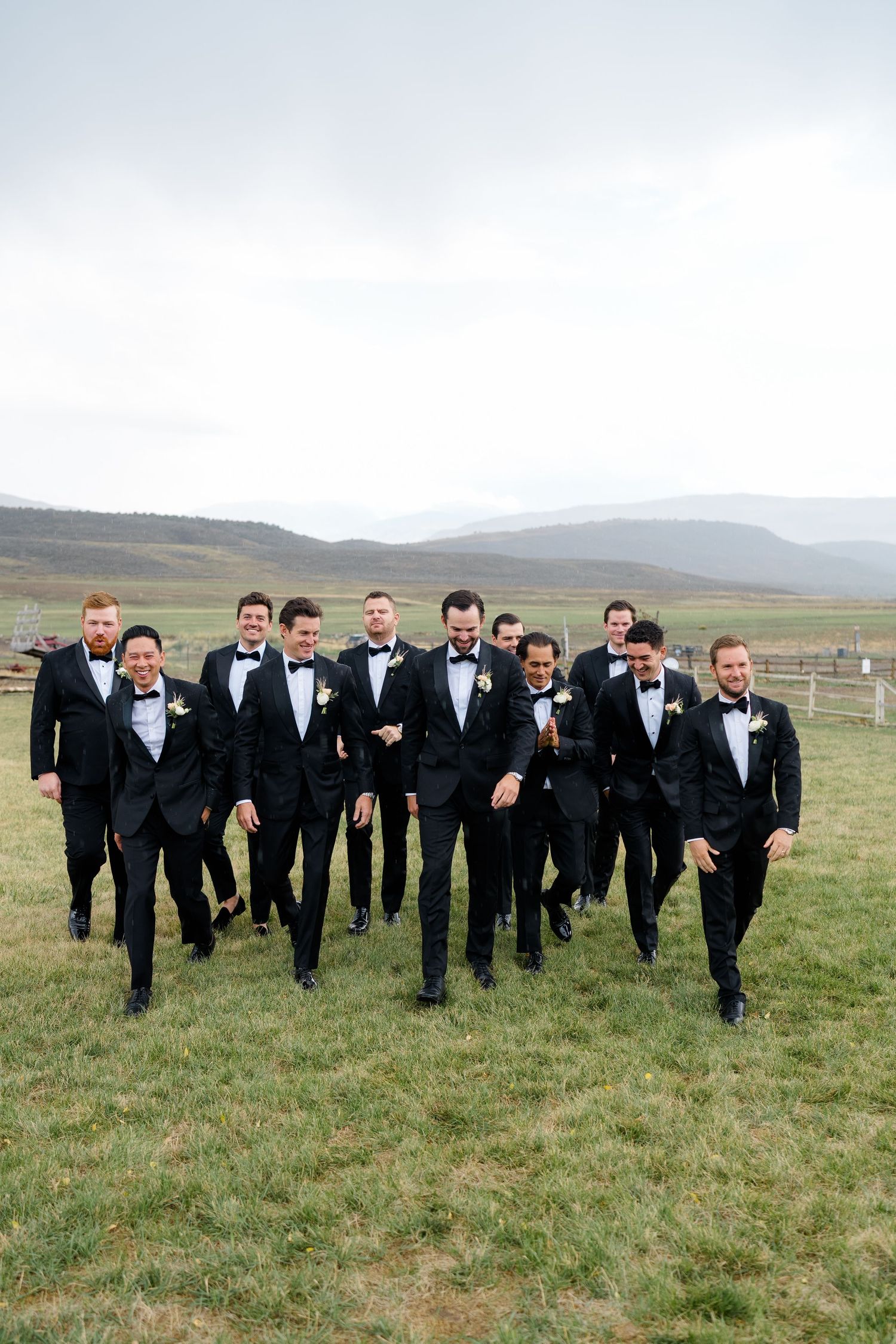 Groomsmen in black tuxedos and bow ties walk together outdoors at a wedding with scenic mountain views.