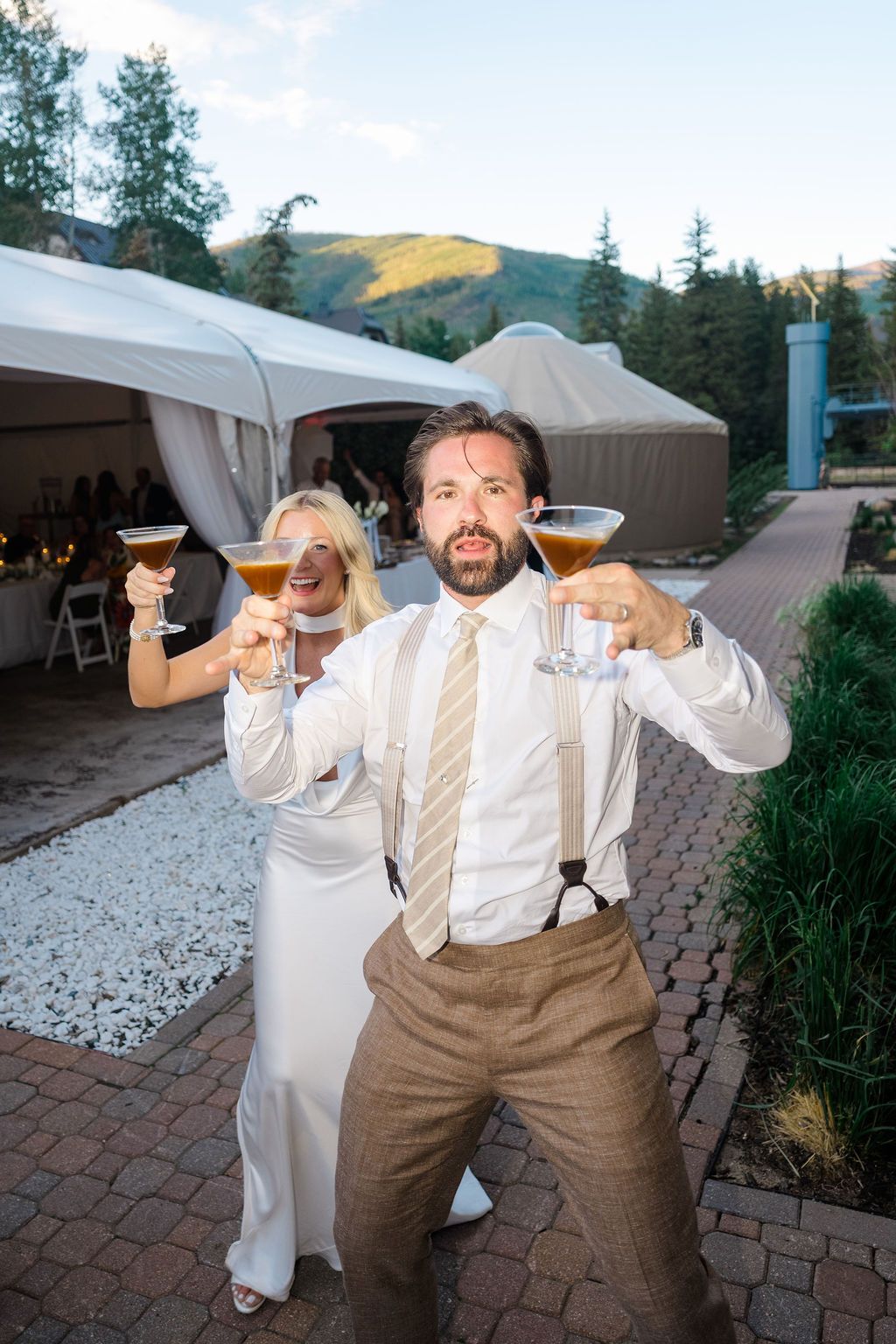 Bride and groom toasting with espresso martinis at outdoor mountain wedding reception near white tent.