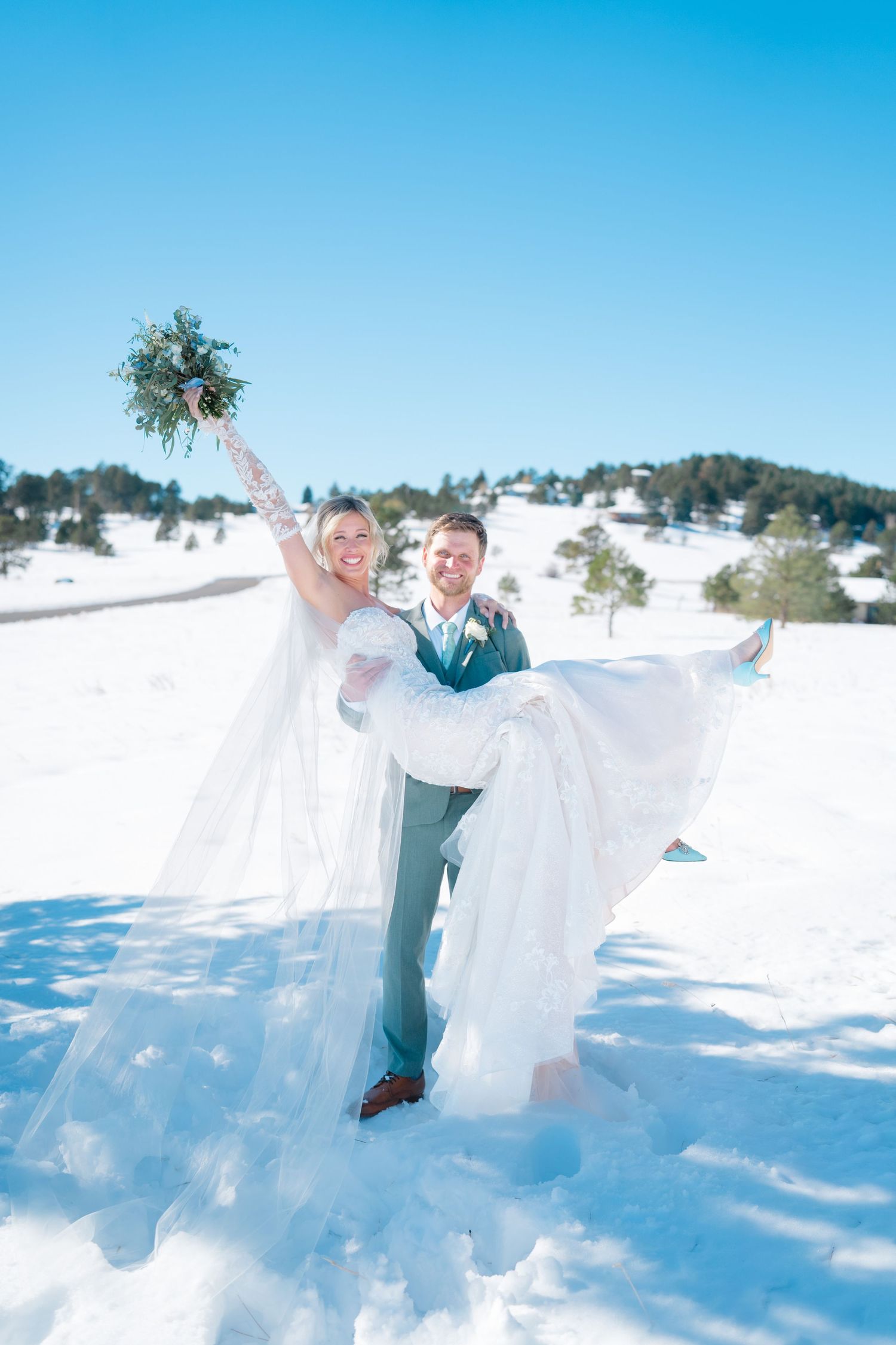 Groom carrying bride in snowy winter wedding, bride holding bouquet aloft, blue sky and pine trees backdrop.