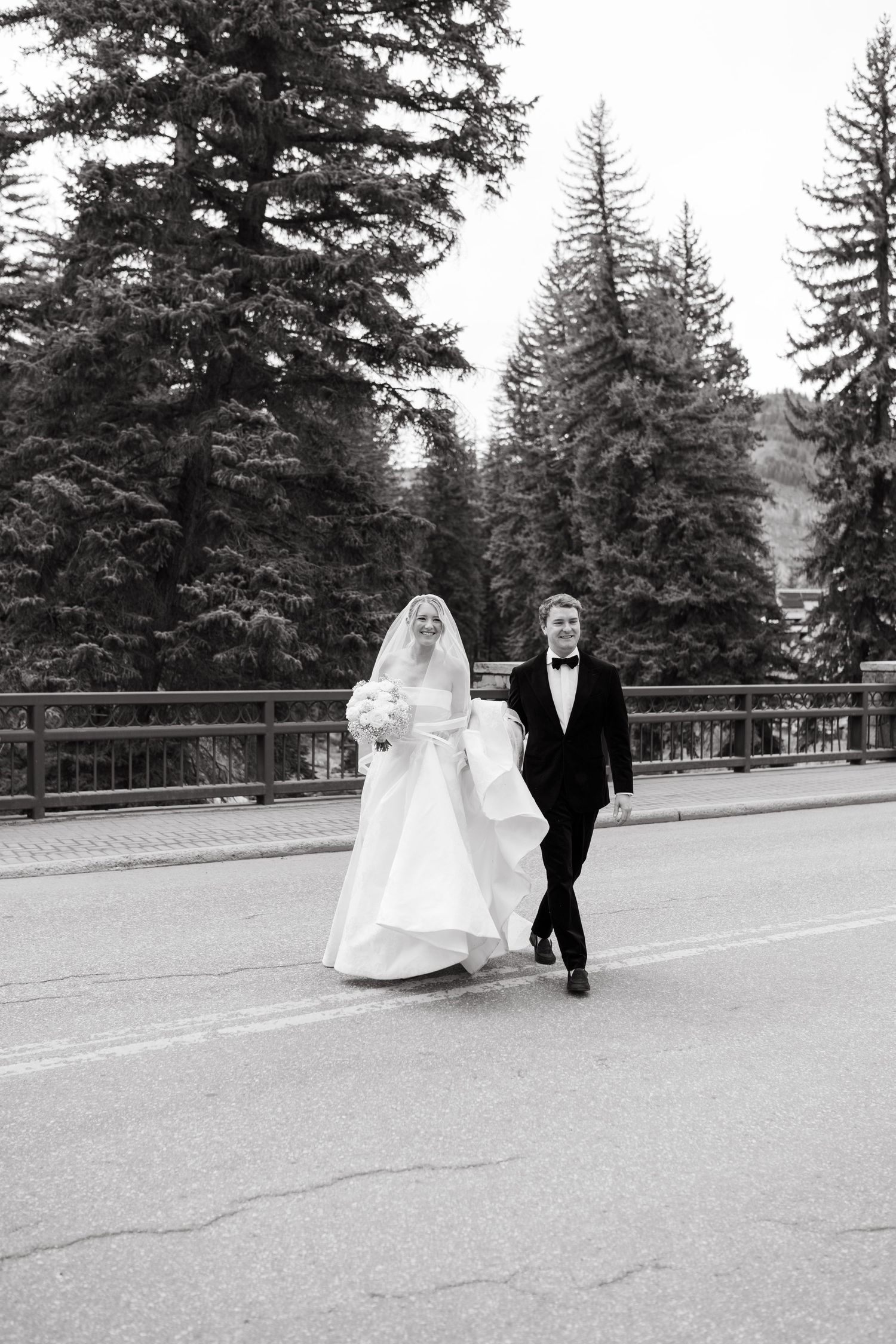 Black and white photo of bride and groom walking hand in hand on a road surrounded by tall pine trees at Vail Sonnenalp Wedding.