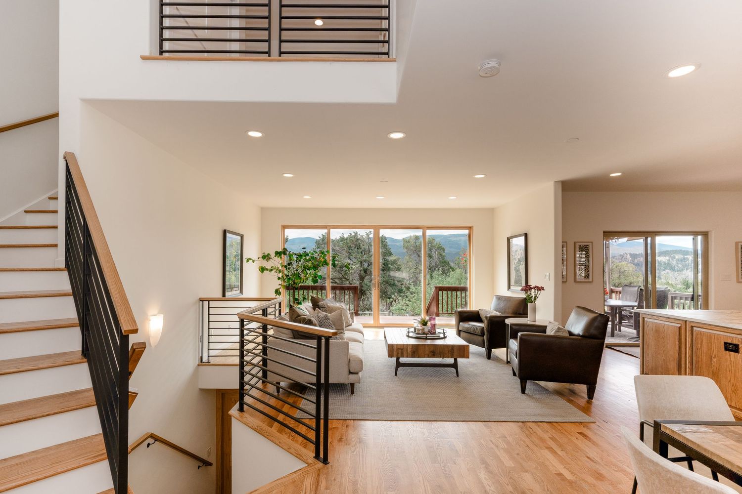 Modern open-concept living room with hardwood floors, high ceilings, staircase, and large windows overlooking lush greenery.