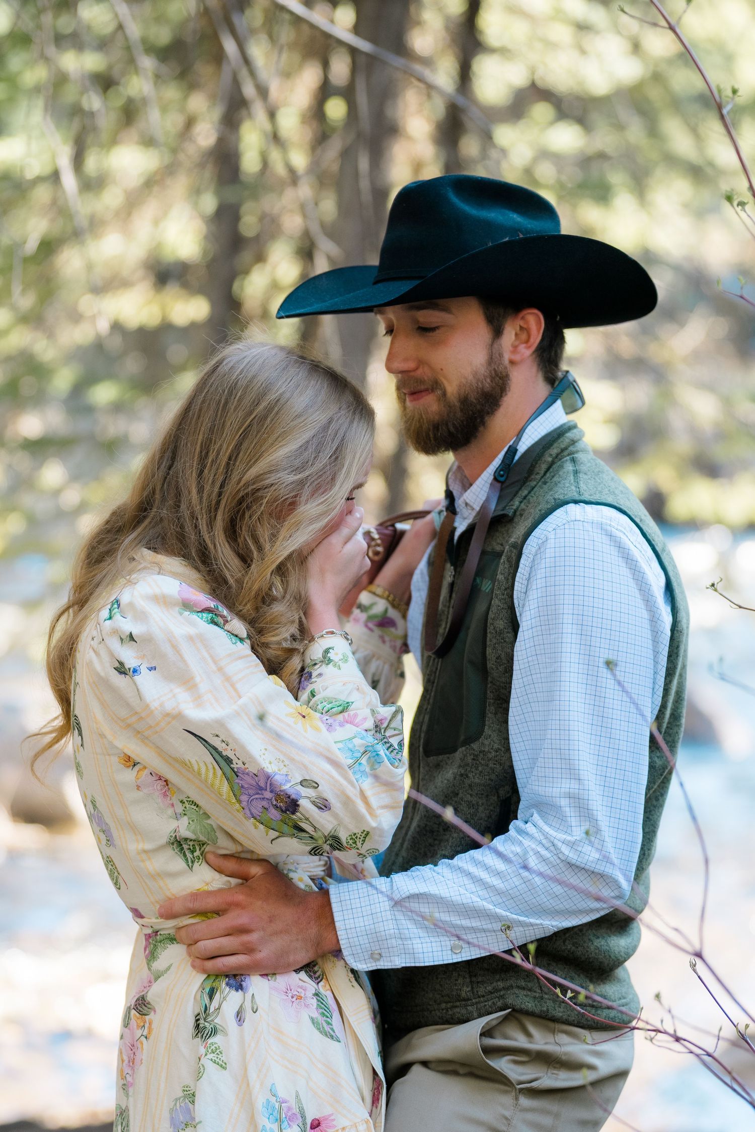 A couple in western attire embraces in a forest, man in cowboy hat and vest, woman in floral dress.