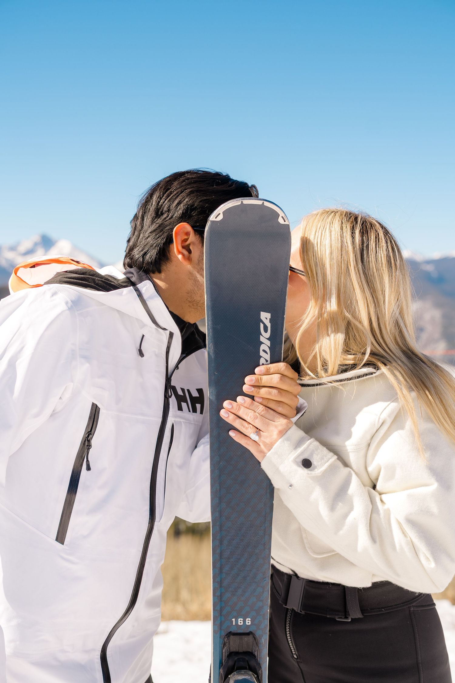 Couple kissing behind Nordica ski on a sunny snow-covered mountain slope.