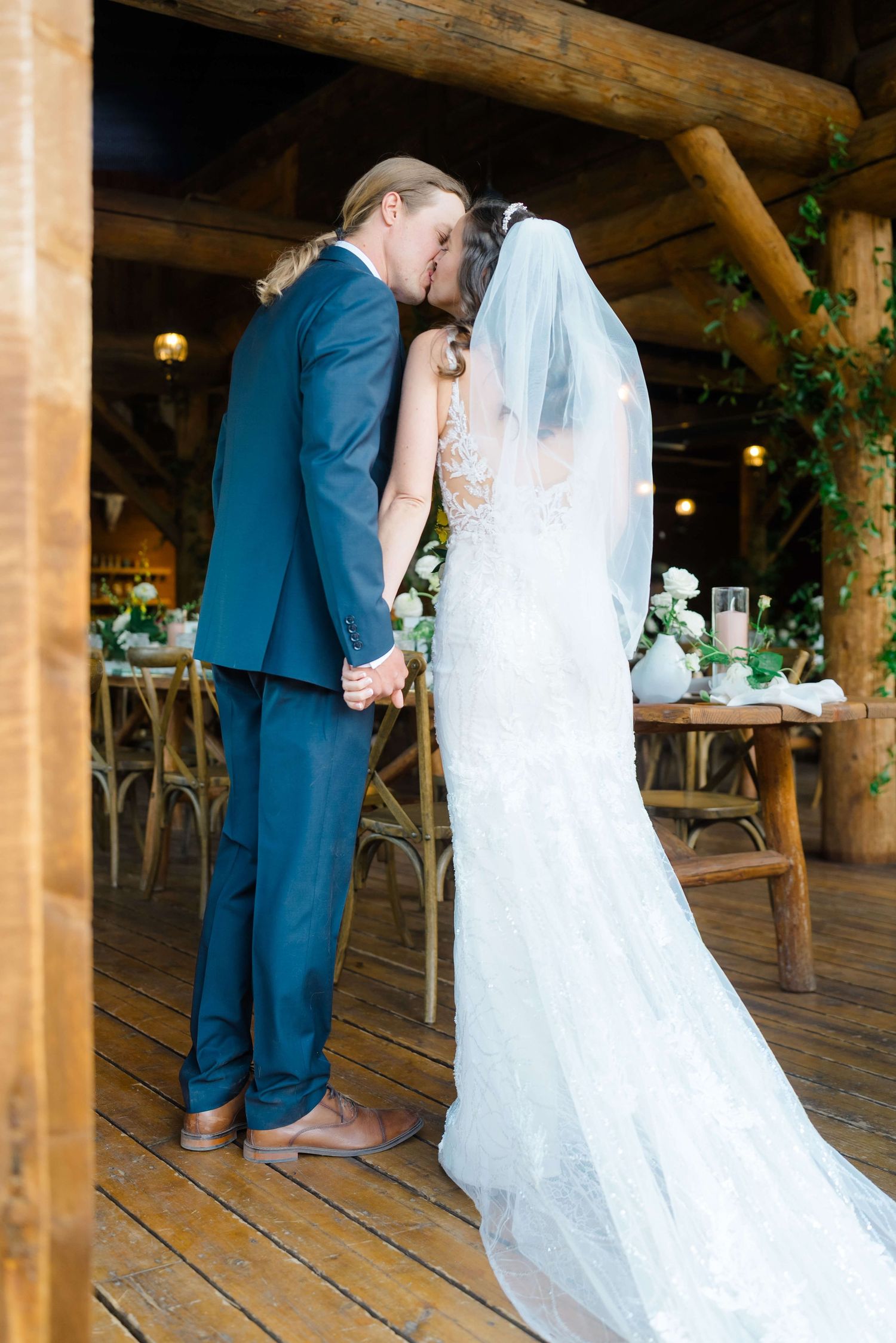 Bride and groom share their first kiss at a rustic barn wedding venue with wooden beams and floral decorations at Piney River Ranch Wedding.