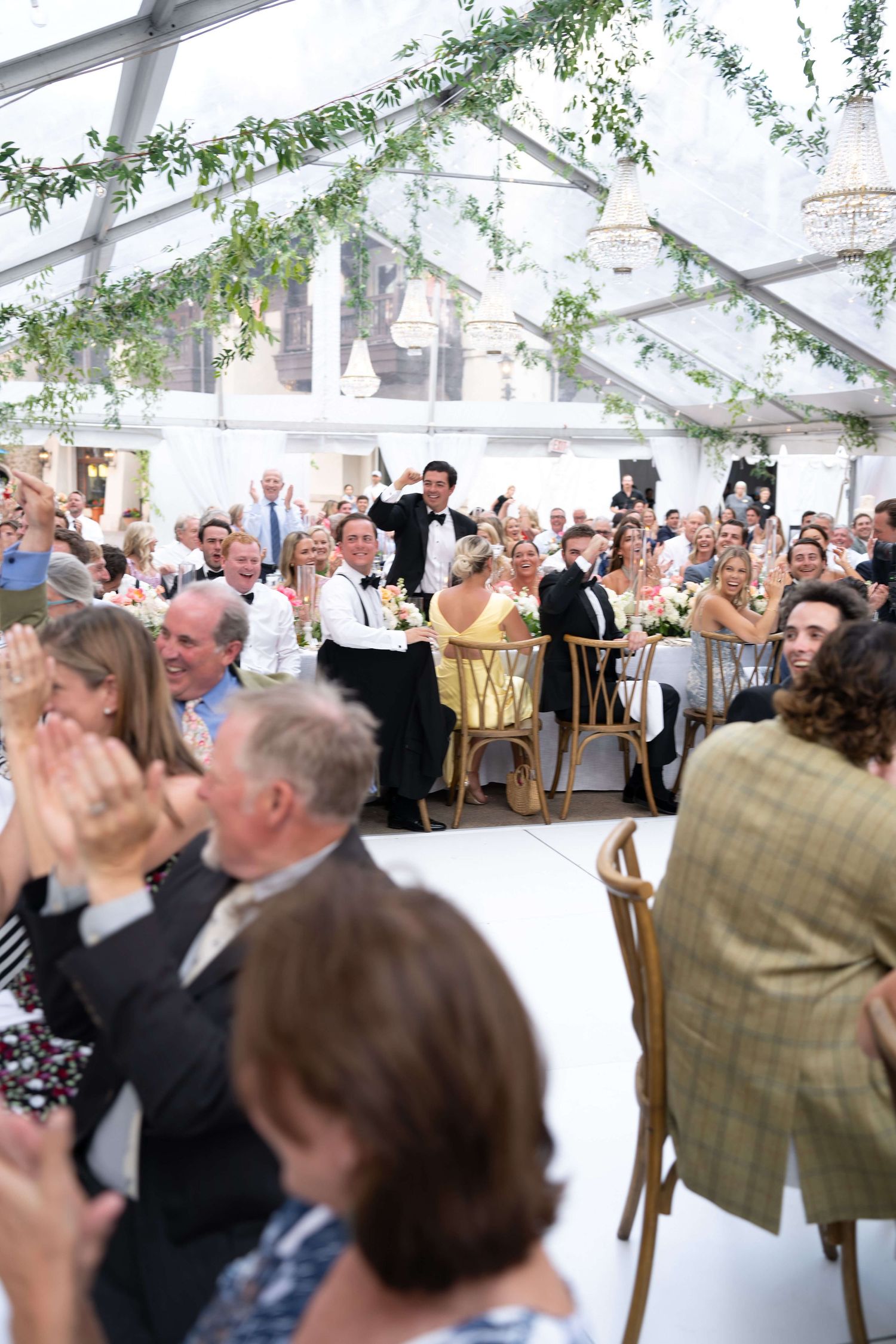 Arrabelle Vail Wedding-Guests clapping and celebrating at an elegant wedding reception under a clear marquee tent decorated with greenery.