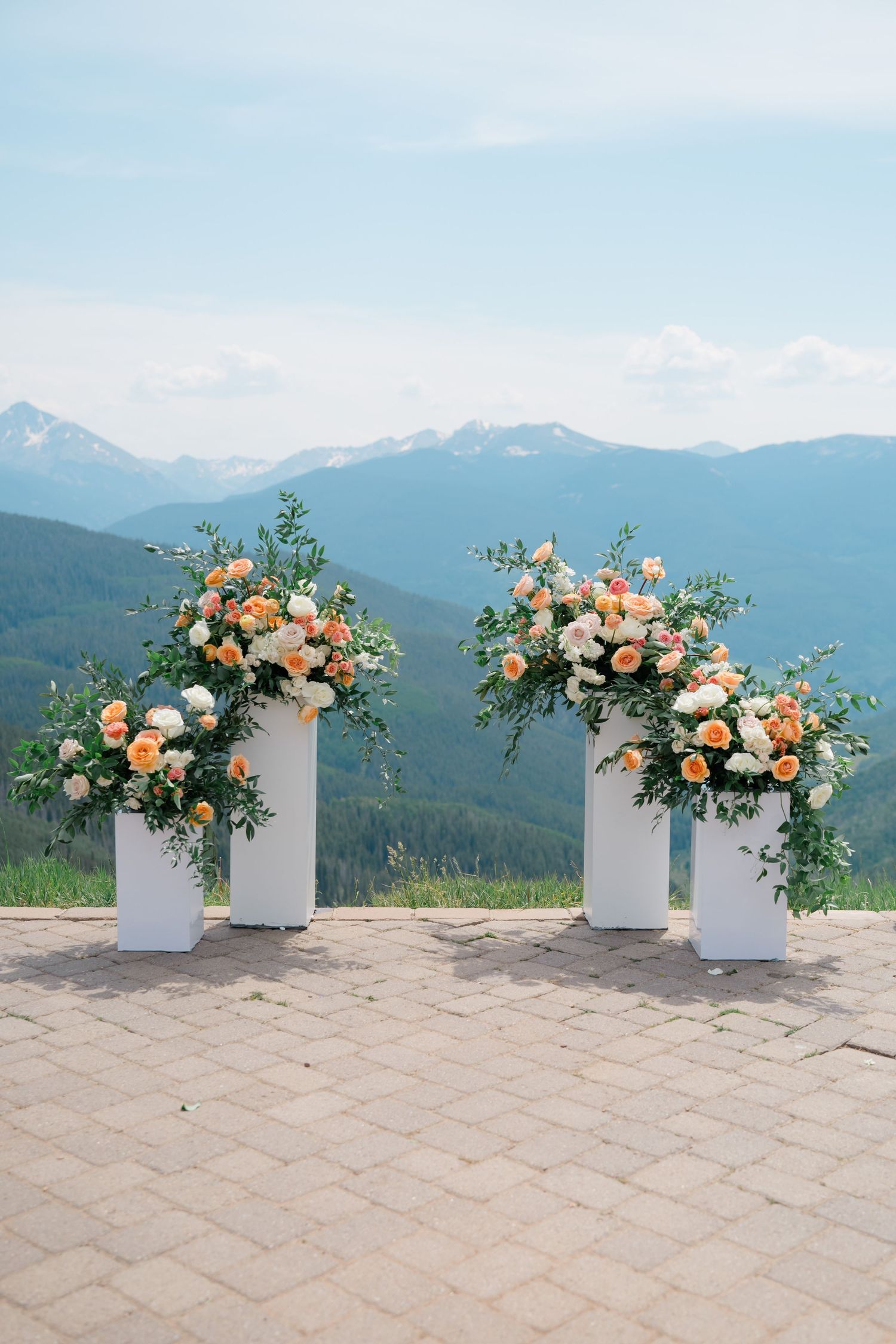 White pedestal floral arrangements with peach and white roses against a stunning mountain backdrop at Vail Wedding Deck.