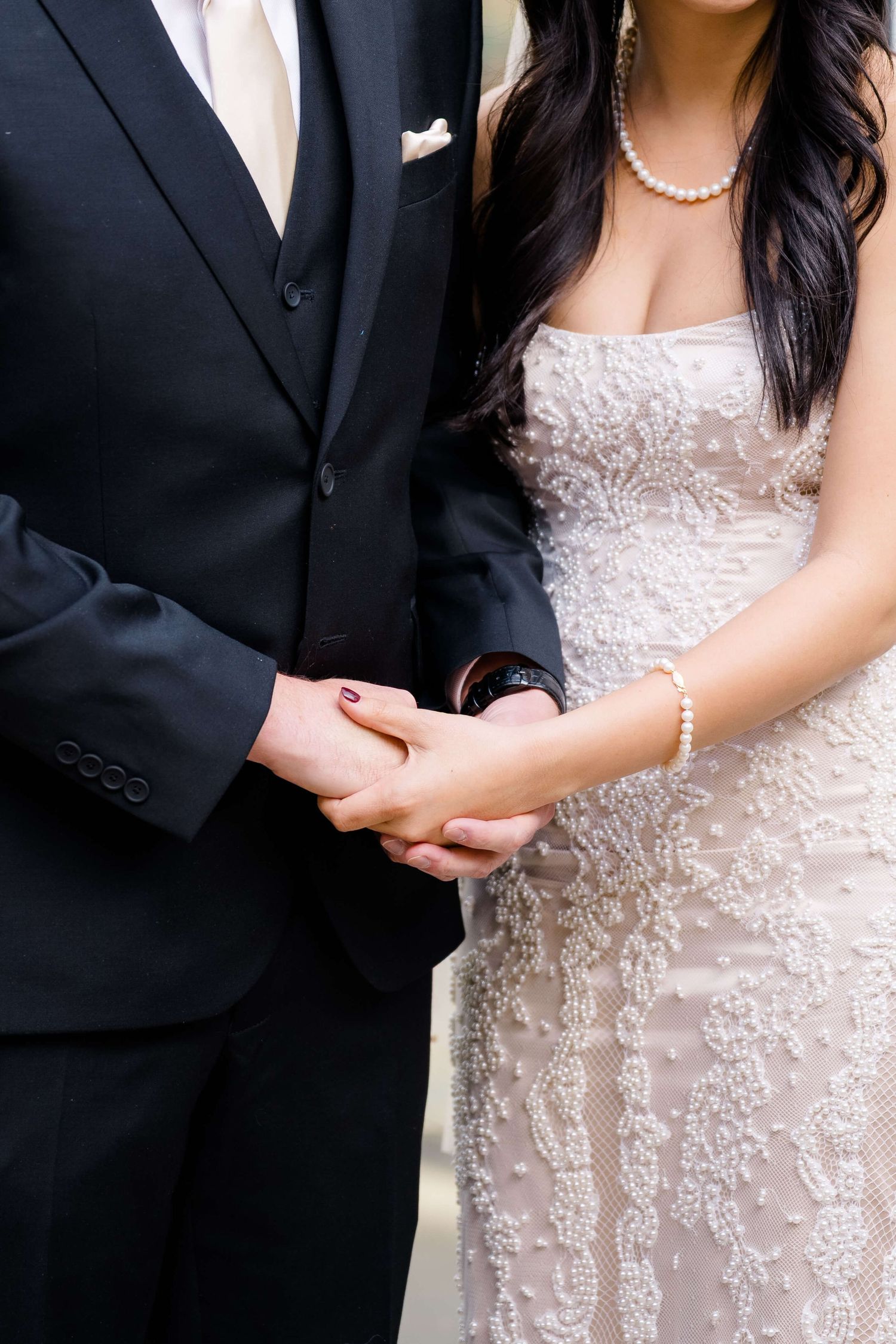 Bride and groom holding hands at wedding, bride in lace strapless gown with pearl jewelry.