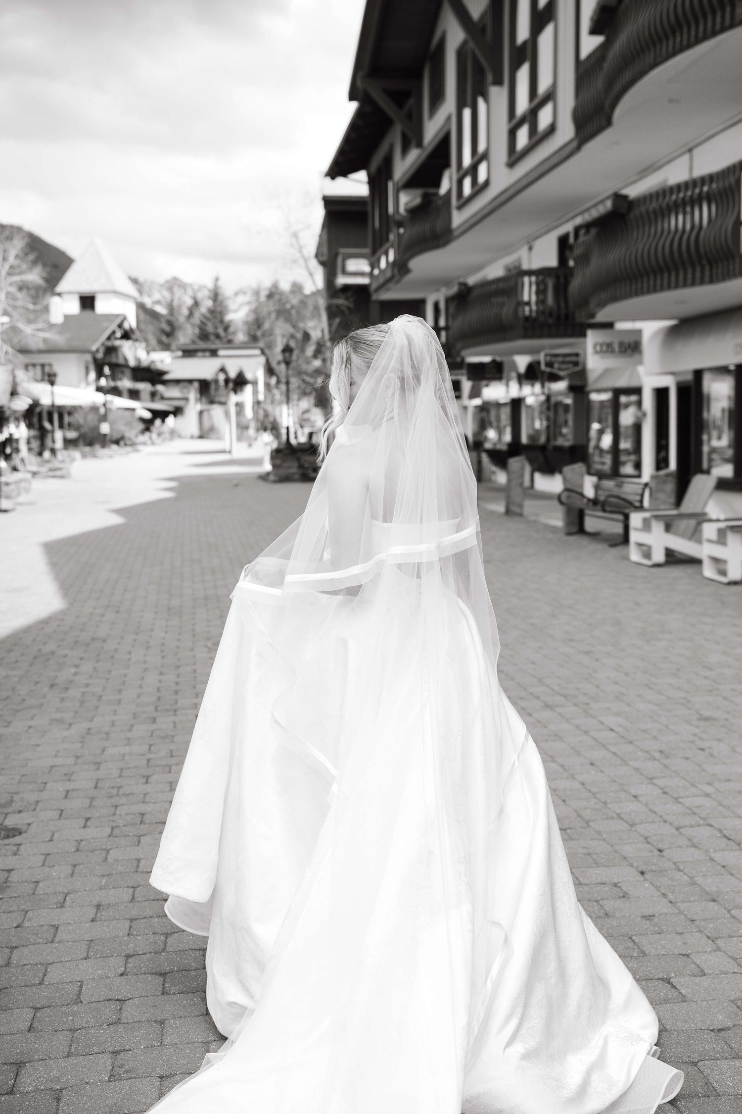 Black and white photo of a bride in a flowing white gown and veil walking down a charming cobblestone street.