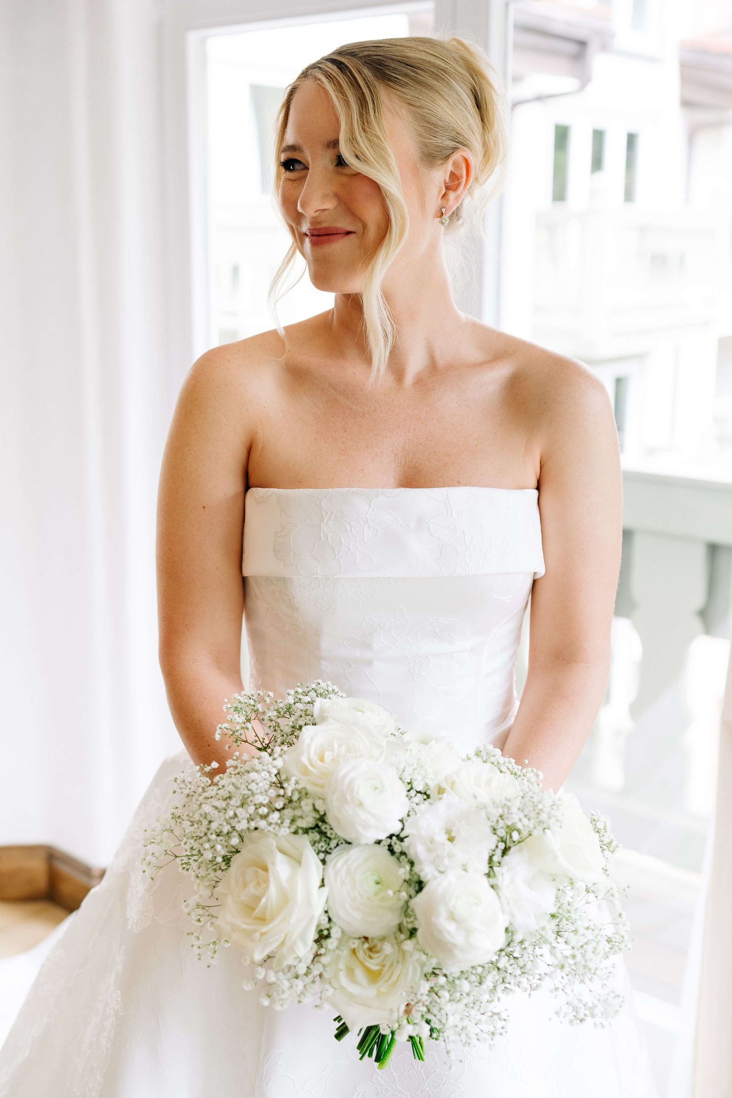 Bride in strapless white gown holding bouquet of white roses and baby's breath, smiling near window.