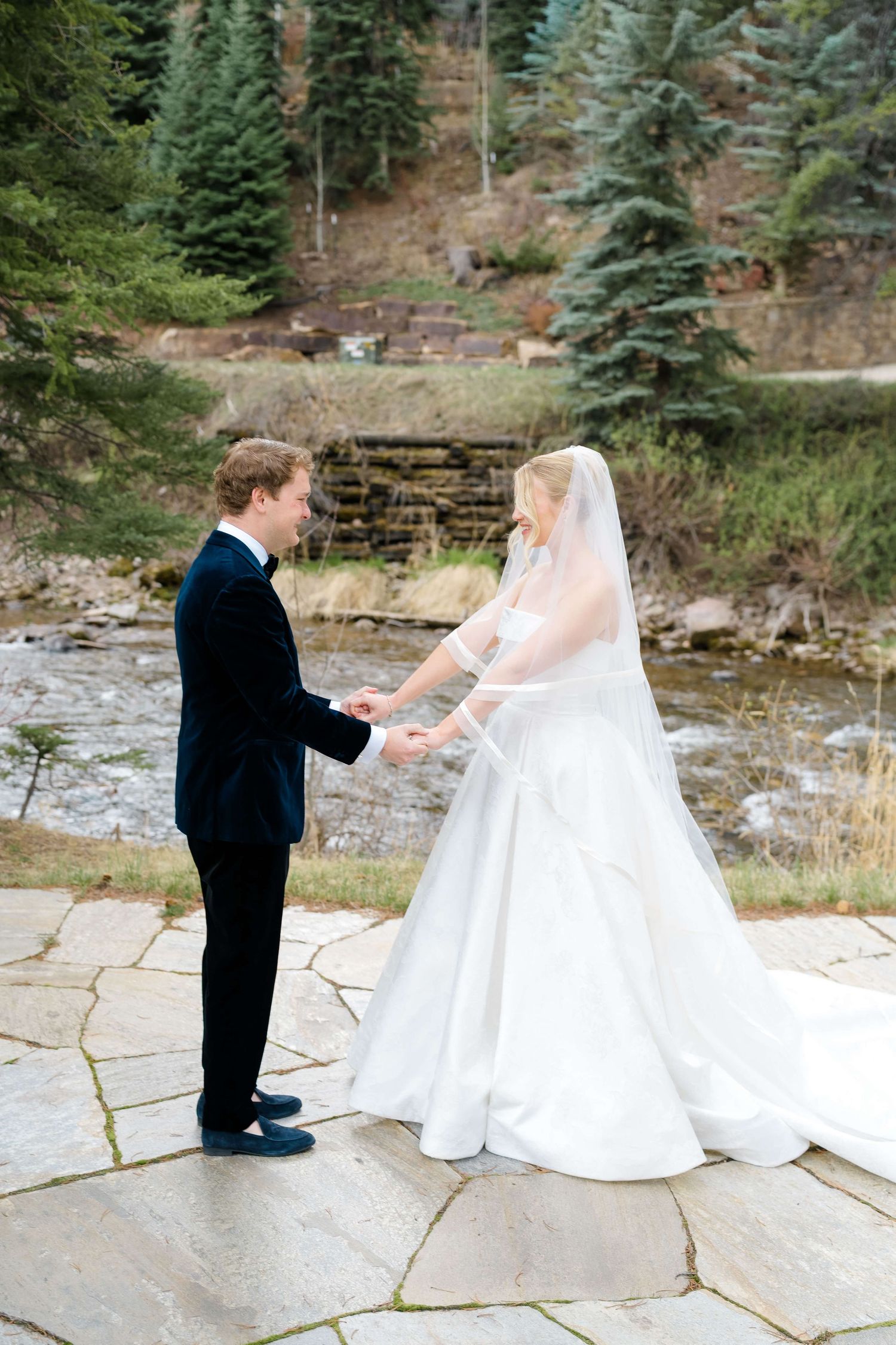 A bride and groom hold hands on a stone patio with a mountain stream and evergreen trees behind them.
