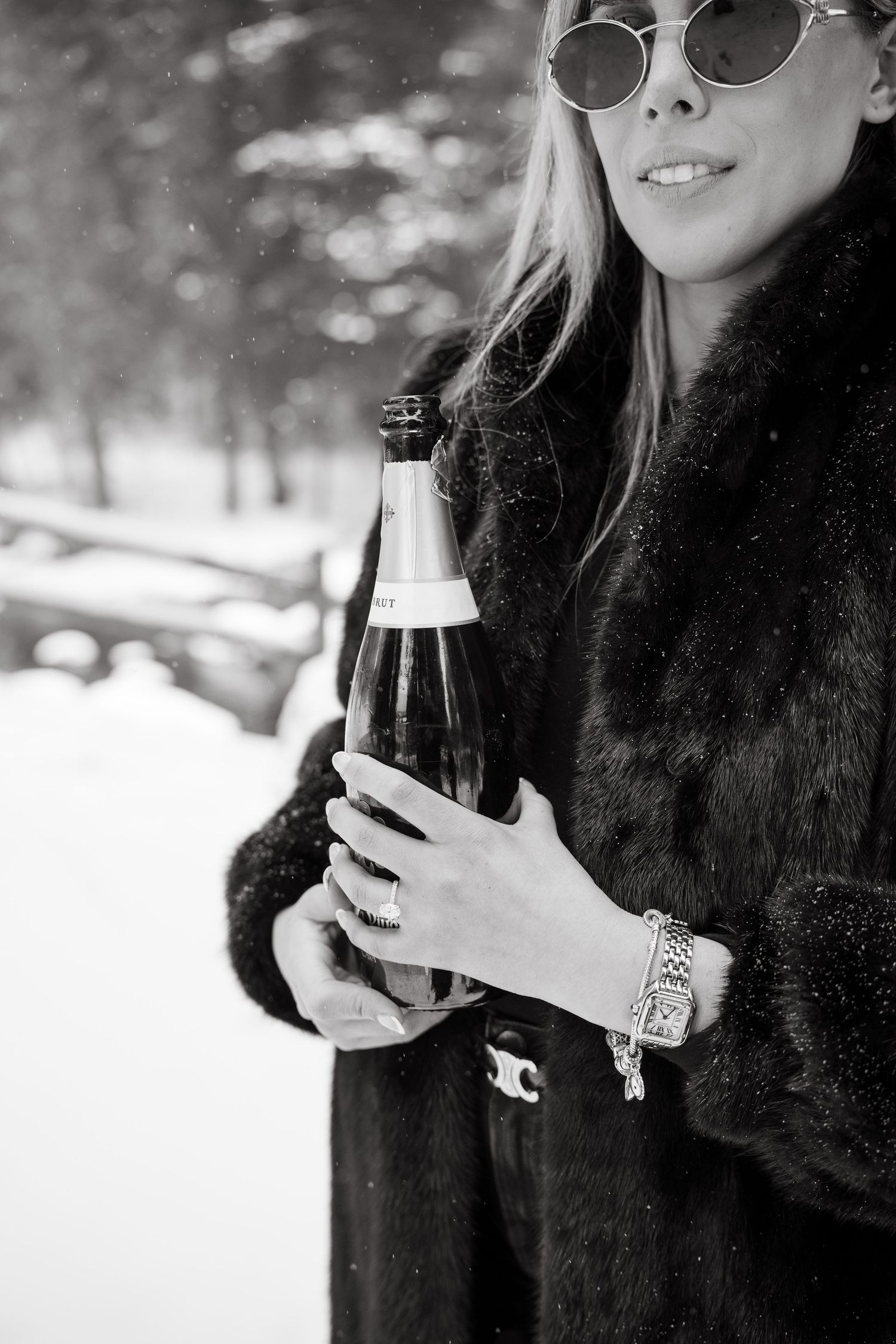 Black and white photo of a blonde woman in fur coat holding champagne bottle outdoors in snowy winter setting.