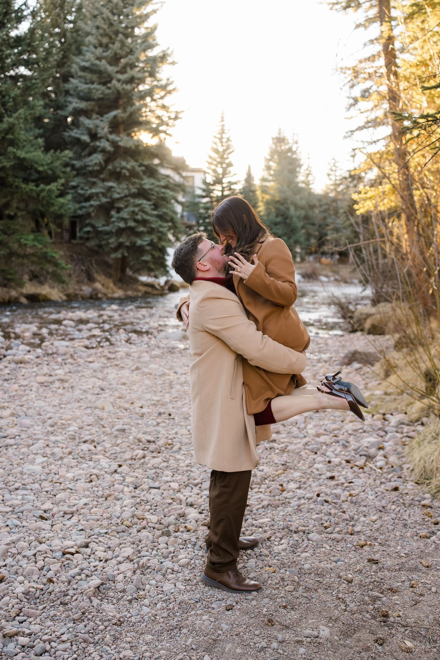 Couple embracing outdoors on rocky path, man lifting woman in matching tan coats, surrounded by pine trees at golden hour.