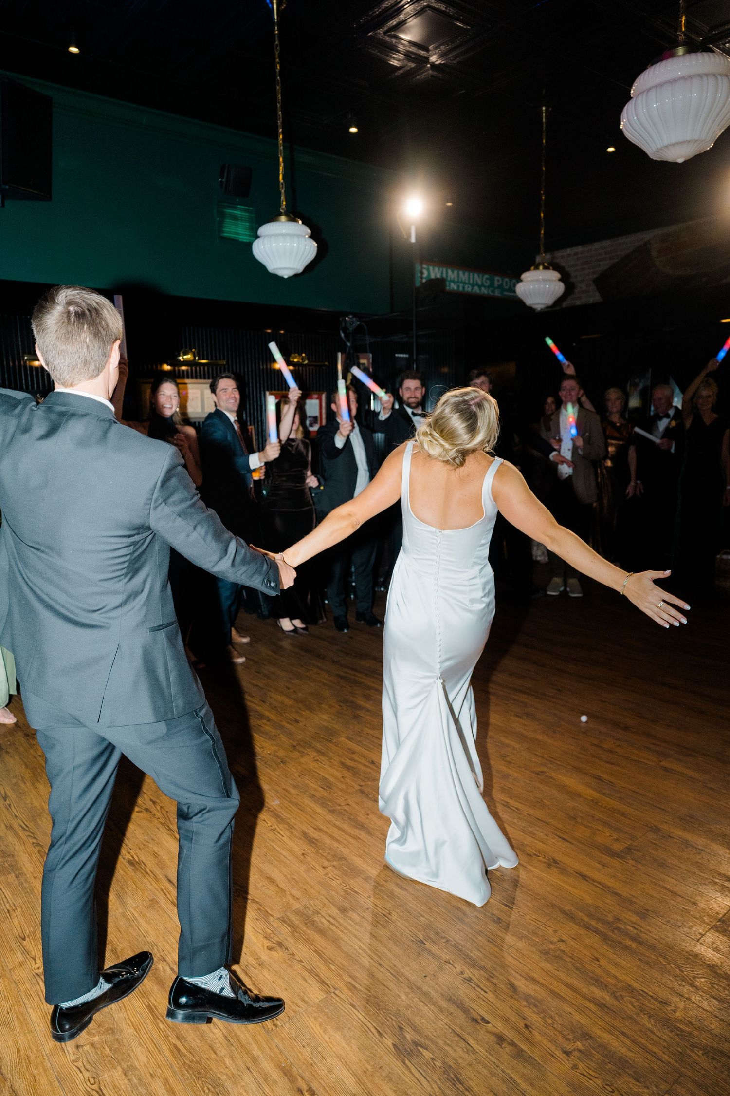 Wedding party dancing on a wooden dance floor under hanging lights at a reception.