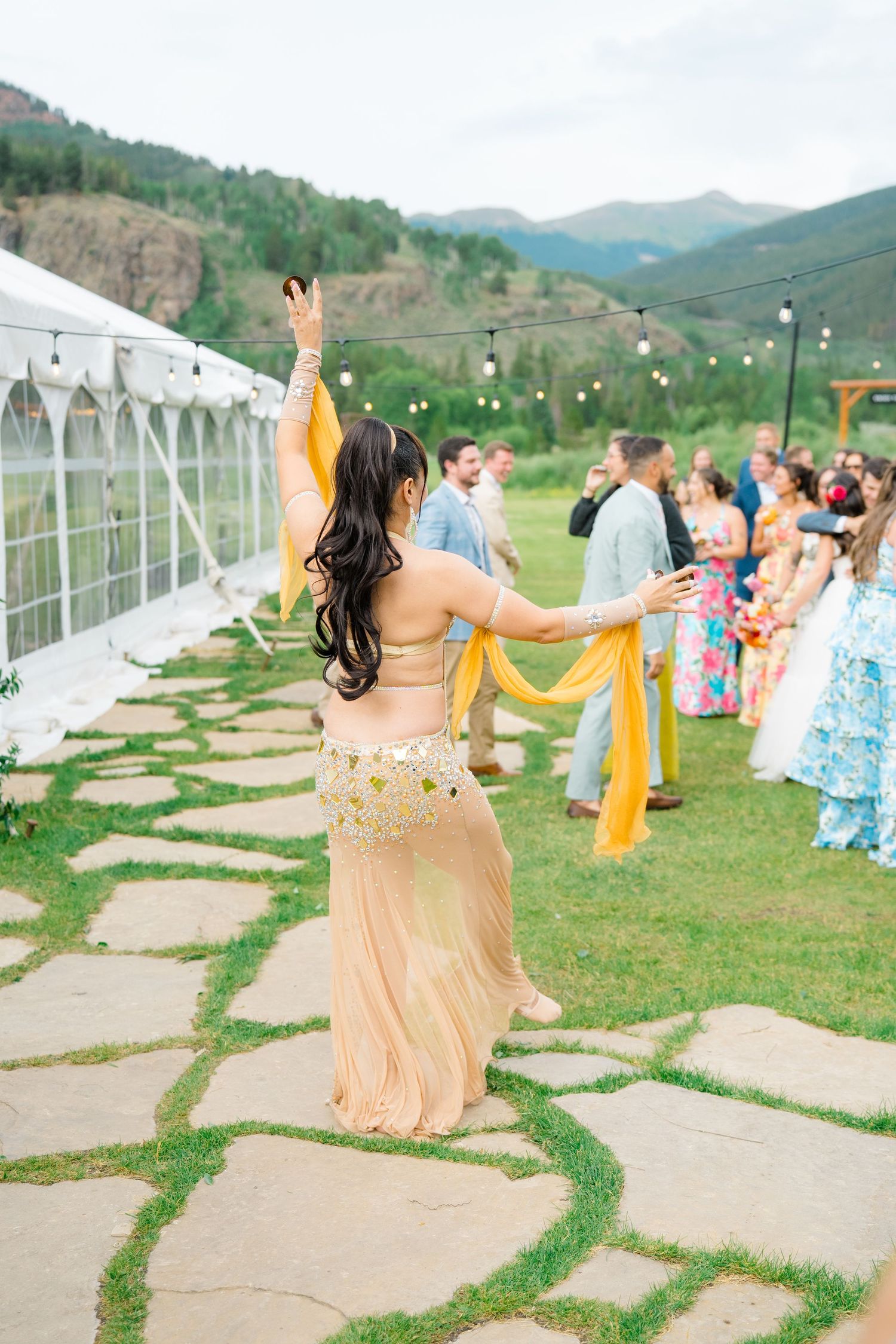 Performer in gold dress dancing with scarves on outdoor stone patio with mountain scenery.