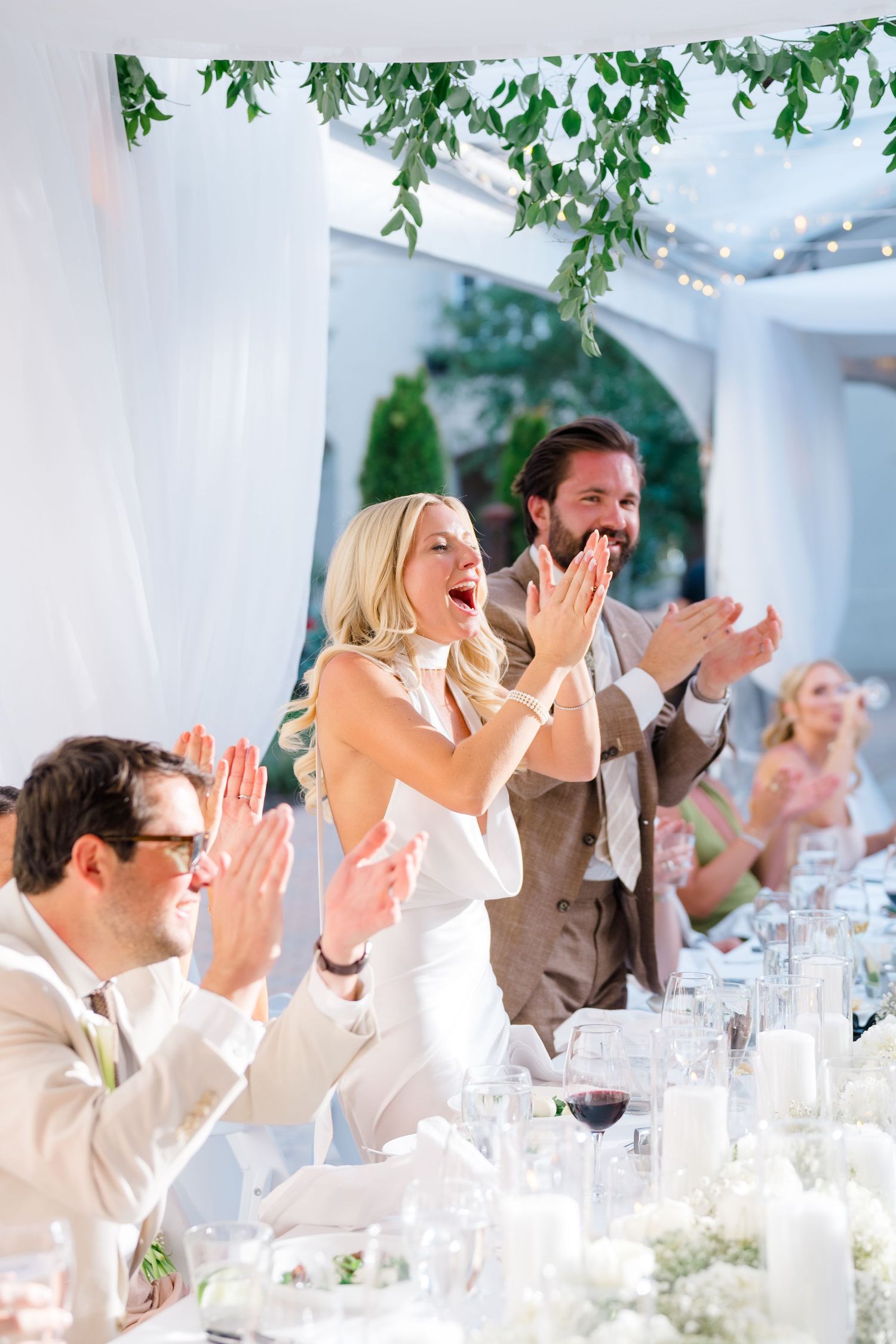 Bride and groom clapping joyfully at elegant outdoor wedding reception with floral decor and guests.
