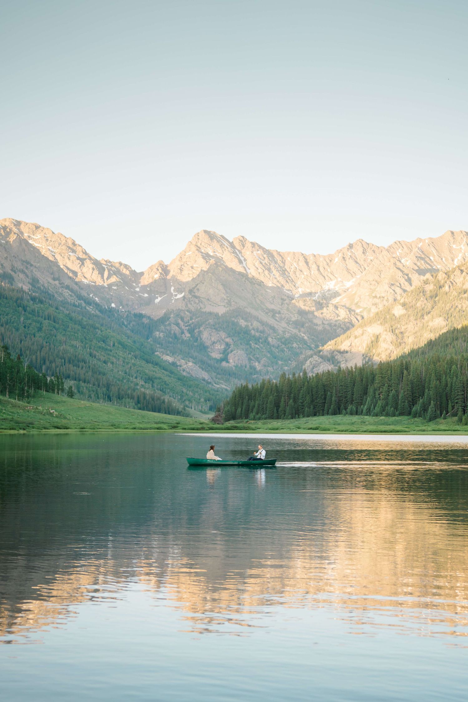 Piney River Ranch Vail Wedding Venue -Two people canoeing on a calm mountain lake surrounded by pine forests and rocky peaks at golden hour.