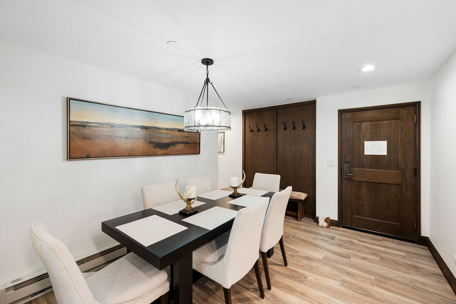 Modern dining room with dark wood table, white upholstered chairs, chandelier, and hardwood floors near a dark wood door.