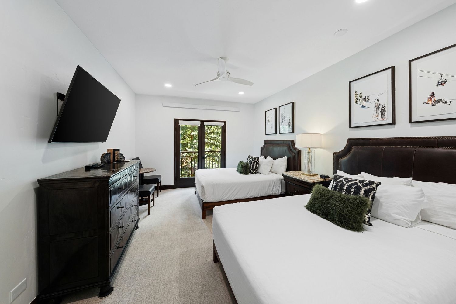 Bright hotel-style bedroom with two queen beds, dark wood furniture, wall-mounted TV, ceiling fan, and French doors to a balcony.