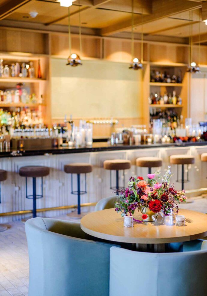 Elegant hotel bar lounge with blue velvet chairs, floral centerpiece, and a well-stocked bar with pendant lights.