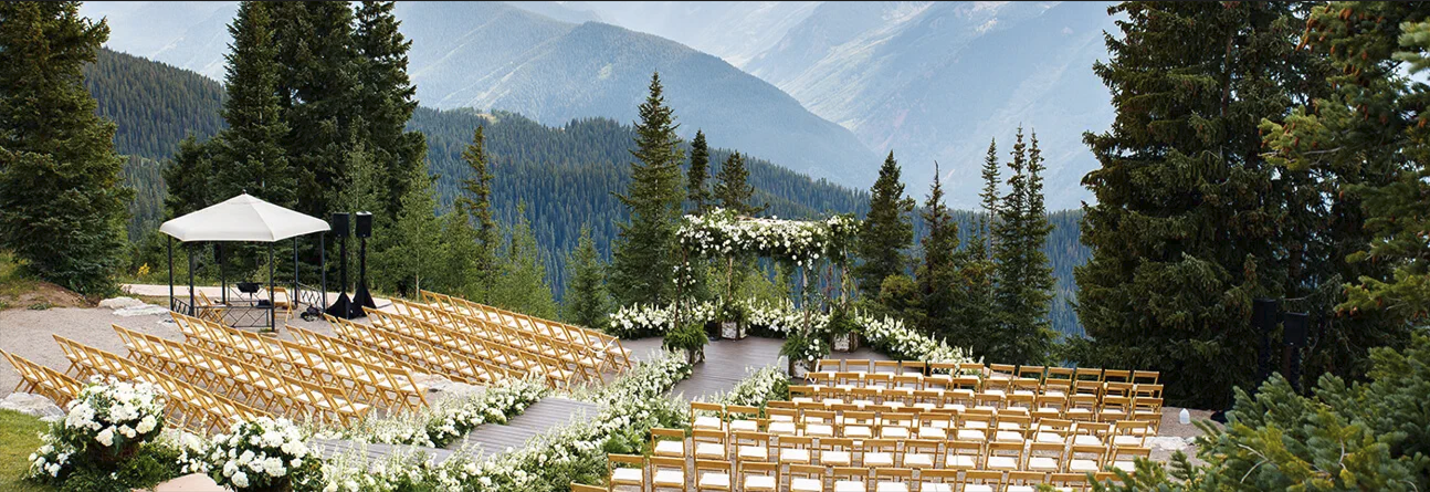 Outdoor mountain wedding ceremony setup with rows of wooden chairs, floral arch, and stunning alpine forest backdrop.