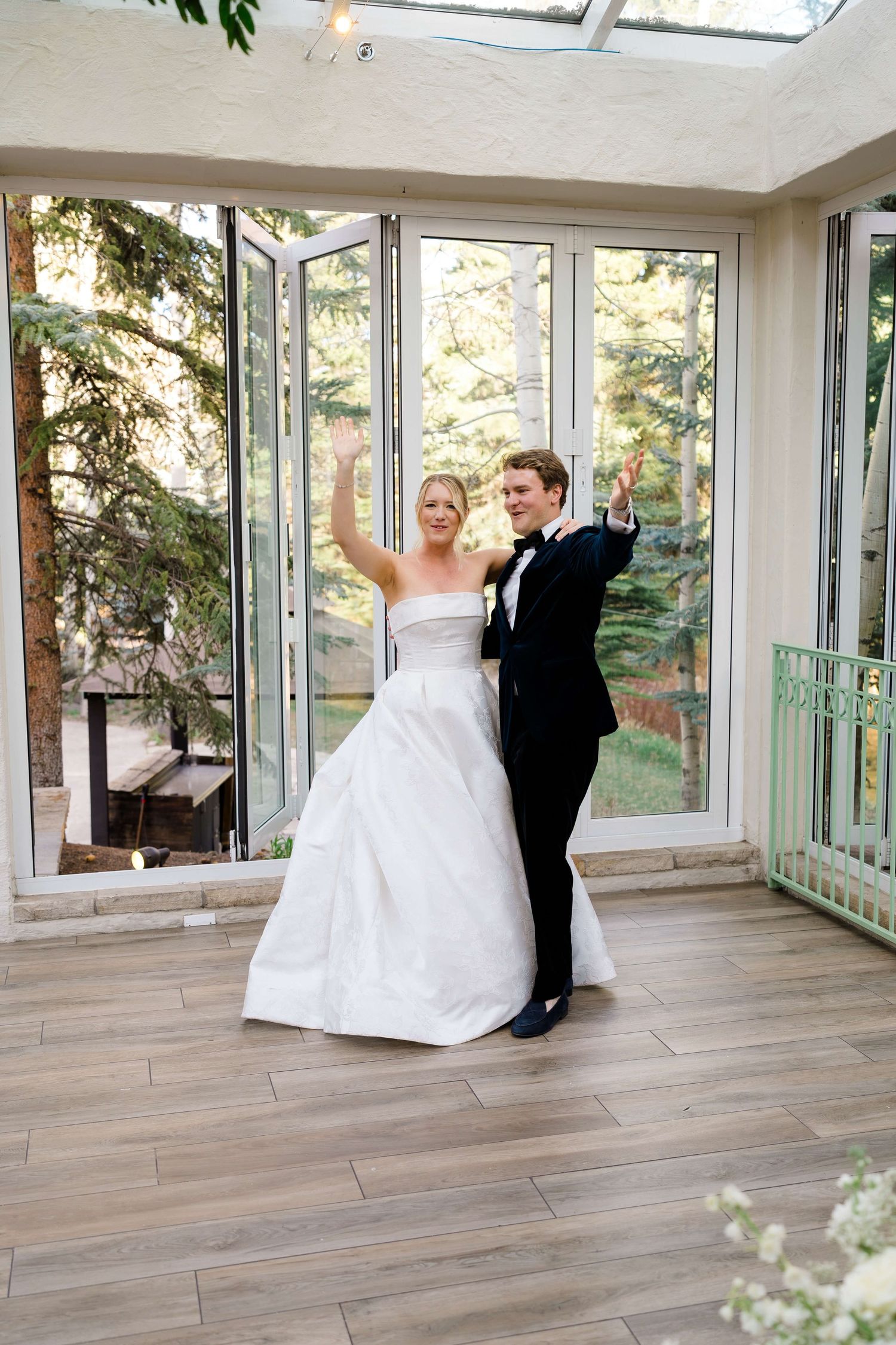 Bride in white gown and groom in black tuxedo raising hands joyfully in a sunlit conservatory.